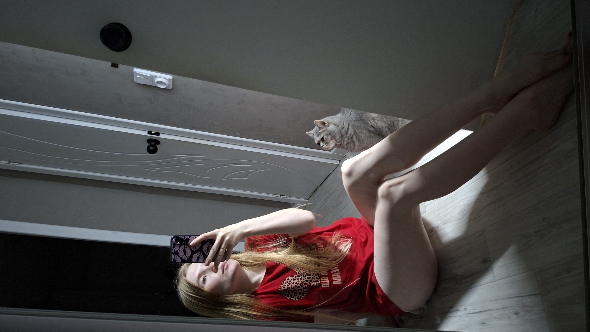 In a dimly lit room, a young woman with long blonde hair sits on a light-colored wooden floor, taking a mirror selfie with her phone. She is wearing a red t-shirt with a leopard print heart graphic. A grey cat sits behind her, looking upwards. The background reveals a white interior door with a dark doorknob and a light switch on the wall. The floor is illuminated by sunlight casting shadows, creating an indoor scene. The location is identified as Chaikovsky, Russia.