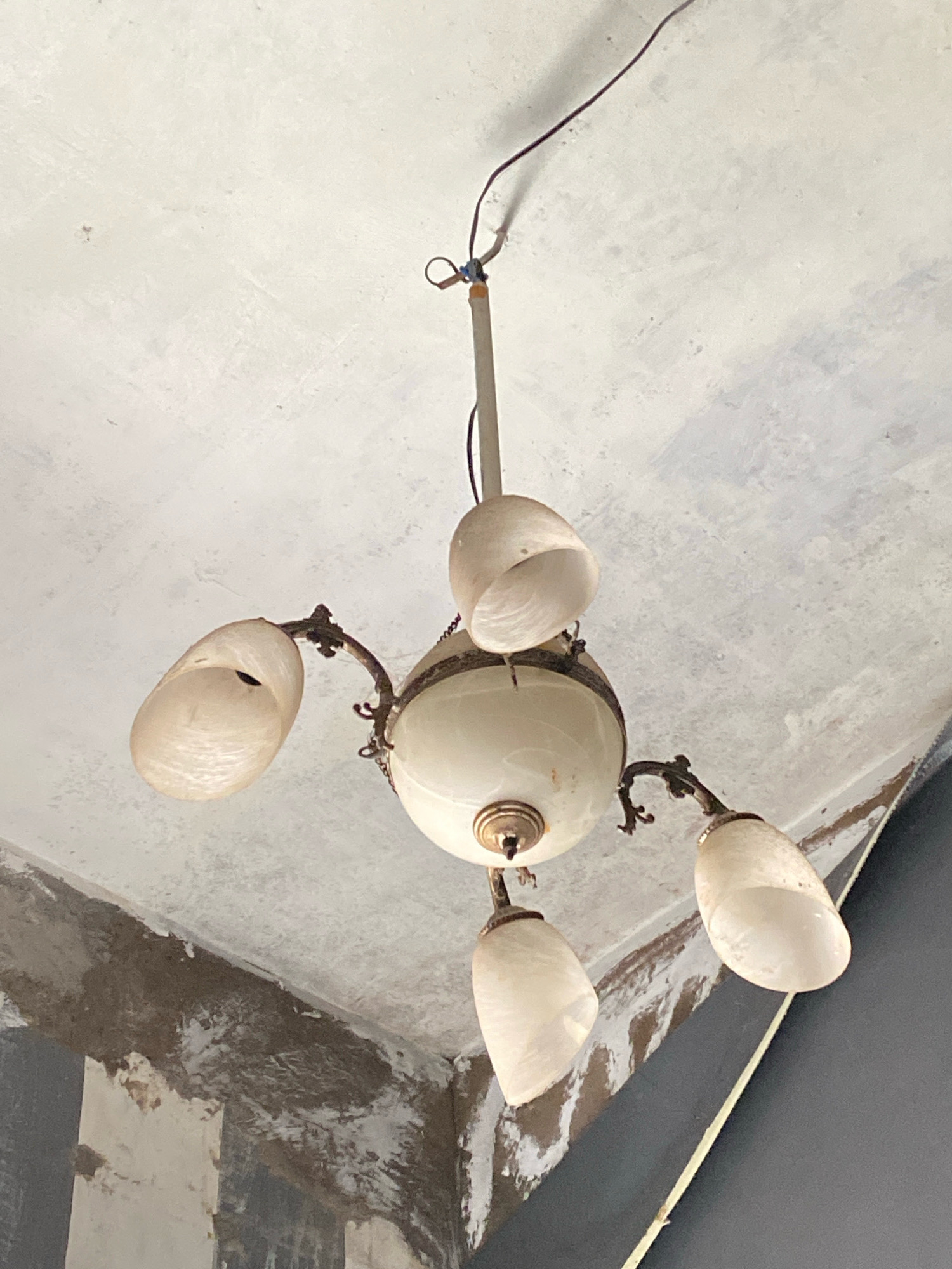 A close-up, low-angle shot captures an antique, cream-colored chandelier hanging from a textured, off-white ceiling. The chandelier has a central body with four arms, each holding a frosted glass shade that appears to be shaped like a conch shell. The shades are translucent, allowing some light to pass through. The chandelier's metal components are a dark, aged brass. The ceiling shows signs of wear and discolored patches, with visible cracks and a rough texture. To the right of the chandelier, a dark gray wall angles upwards, partially obscuring the view. A thin, yellow electrical wire hangs loosely from the ceiling, near the chandelier's suspension point, suggesting it might be disconnected or in disrepair. The overall impression is one of an old, possibly neglected interior space.