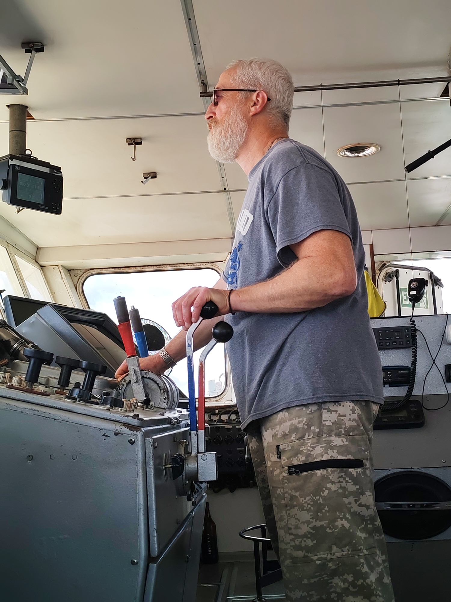 A Caucasian man with a white beard and grey hair, wearing glasses, a grey t-shirt, and camouflage pants, stands in the pilot's cabin of a ship. He is looking to the right and his right hand is on a lever. The cabin is equipped with various instruments, including a control panel with several levers, a monitor mounted on the ceiling, and a radio. Through the window, a hazy sky and distant structures are visible.
