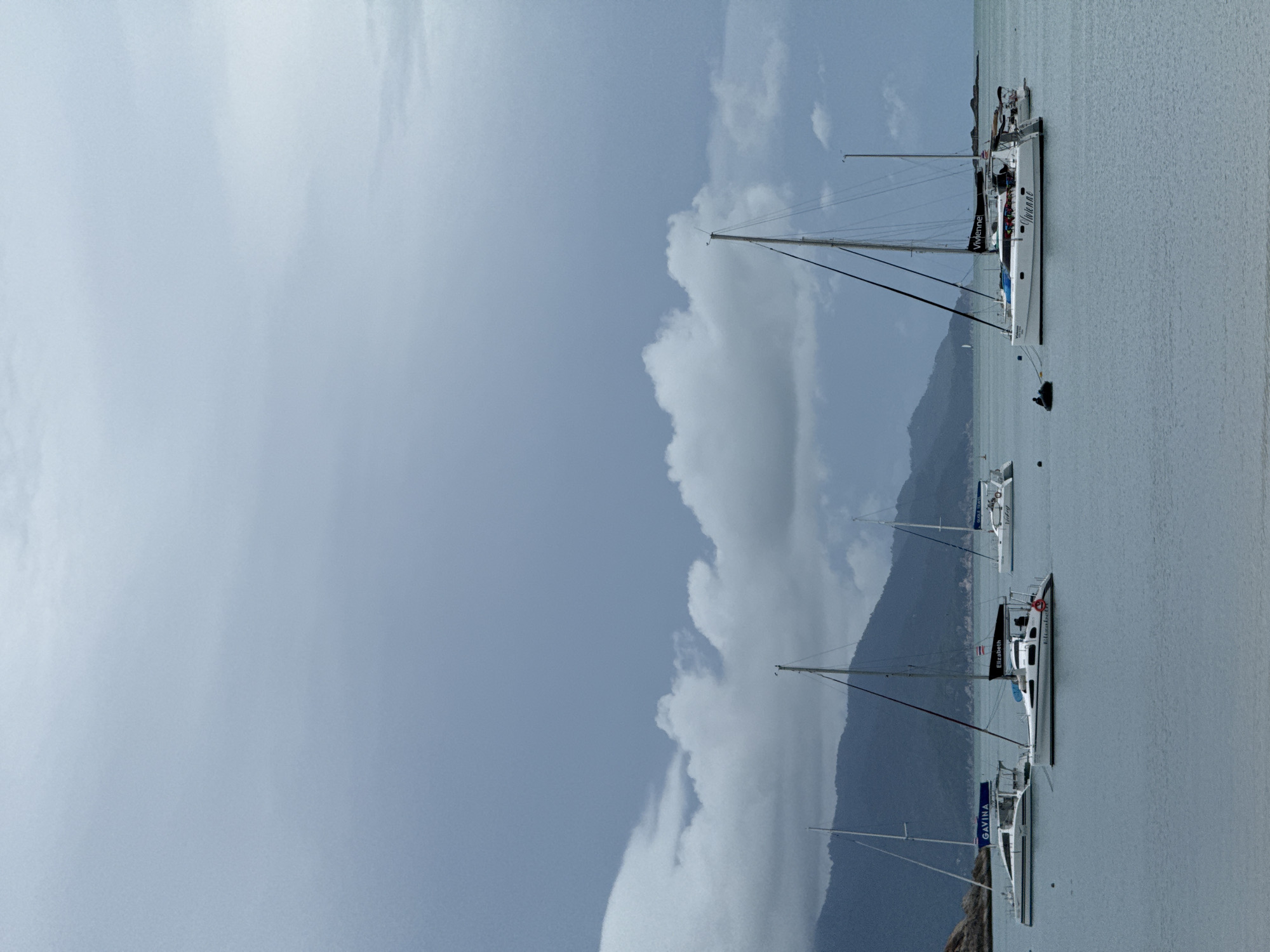 The image depicts a serene bay in Ko Samui District, Thailand, under a cloudy sky. Several white catamarans are anchored in the calm, blue water. One catamaran in the foreground is named "Vivienne" and has a colorful array of items on its deck. Another catamaran to its left is named "Elizabeth." Further in the distance, other catamarans are visible, including one with "Gavina" printed on its canopy, and another with "Vivien" on its side. The background features a lush, green mountain range, characteristic of the island's landscape. A small figure is present in the water near the "Vivienne" catamaran, appearing to be on a small inflatable craft. The overall atmosphere is tranquil, suggesting a peaceful afternoon in a tropical paradise.