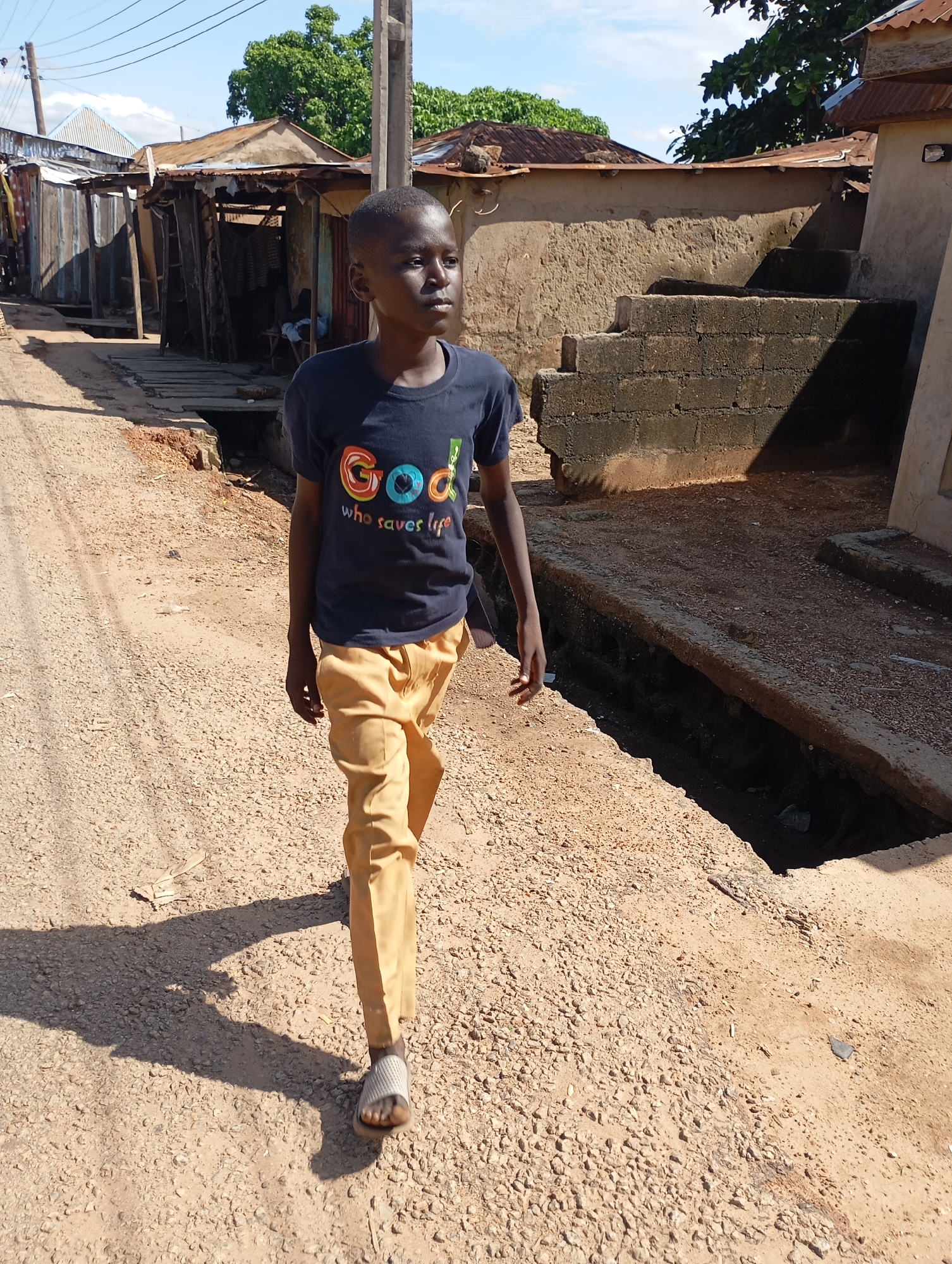 A young boy walks down a gravelly, unpaved road in Shendam, Nigeria. He is wearing a dark blue t-shirt with colorful lettering that reads "God who saves life" and mustard-yellow pants. His gaze is directed to his right, away from the camera. To the left of the road are several makeshift structures with corrugated metal roofs and wooden walls, suggesting a residential or commercial area. To the right of the road, there is a raised concrete structure with a few steps, and a dark, open trench runs alongside it, filled with debris and shadows. The sky above is blue with scattered clouds, and sunlight casts long shadows from the structures and the boy onto the road. The overall scene depicts a typical street environment in a Nigerian town.