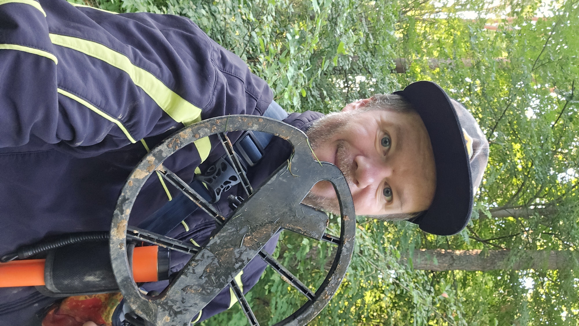 A man wearing a dark blue jacket with yellow accents and a plaid baseball cap is holding a metal detector in front of his face. He is outdoors in a wooded area with green foliage and trees visible in the background. The metal detector's coil is dirty, suggesting it has been used. The man is smiling and looking towards the camera. The scene appears to be in Lobkovo, Russia.
