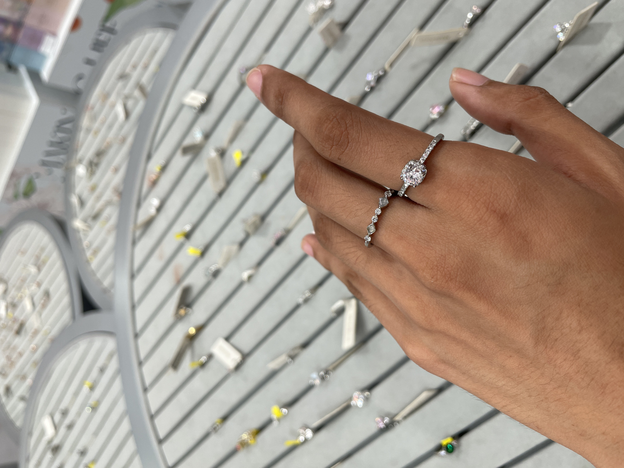 A hand with two rings is positioned over a display of jewelry, likely in a store. The hand is an olive-toned skin tone, with some fine hairs visible on the back. The rings are delicate and silver-toned. The top ring is a band with small, clear stones, and the bottom ring features a larger, round, clear stone surrounded by smaller stones. The jewelry display is grey with angled grooves, and various other pieces of jewelry, many with small price tags, are visible in the background. The lighting suggests an indoor environment. The image was taken in the city of Davao City, Philippines.