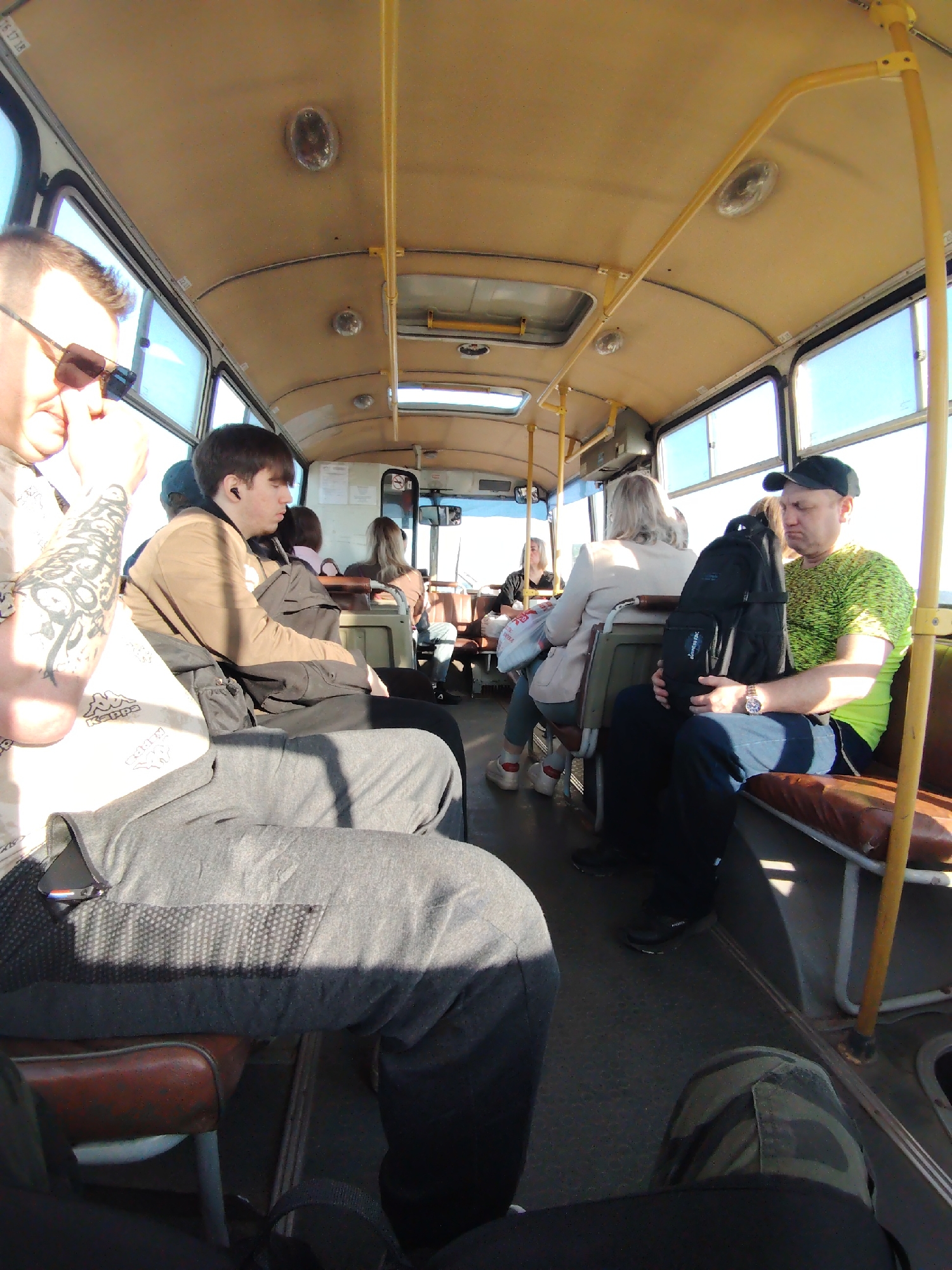 The image depicts the interior of a bus. Several passengers are seated, facing forward. To the left, a man with sunglasses and tattoos on his arm is visible. Next to him is a younger man with an earbud. Further back, other passengers are present, including a woman in a light-colored jacket and a man in a green shirt and black cap holding a backpack. The bus has a yellow ceiling and handrails, with windows along the sides providing natural light. The seats are upholstered in brown. The location is assumed to be a bus in Chaikovsky, Russia based on the content of the image.