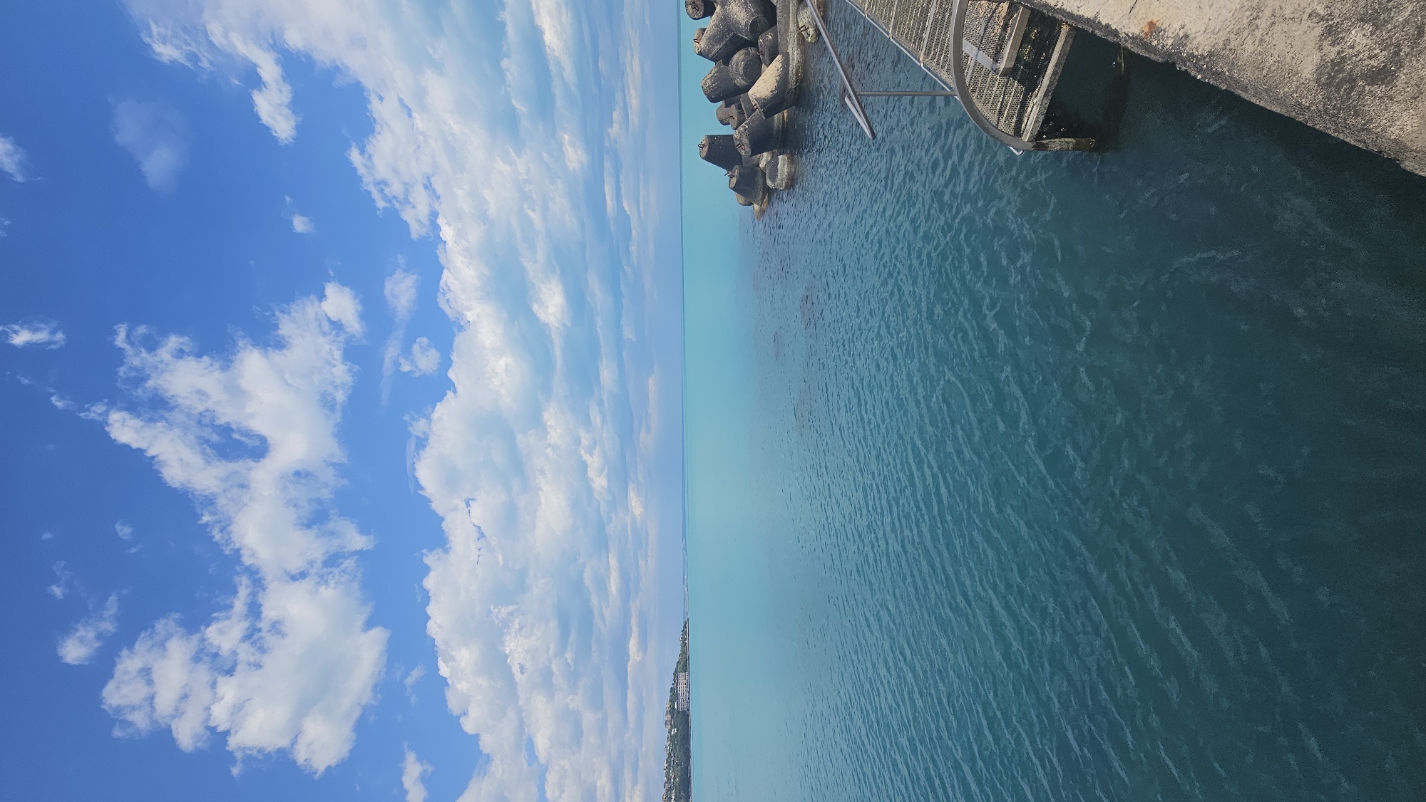The image shows a view of the Black Sea in Varna, Bulgaria. The sky is predominantly blue with scattered white clouds. The sea is a gradient of turquoise near the horizon to darker blue closer to the foreground. Concrete tetrapods are visible along the coastline near a concrete pier with a metal ladder leading into the water. A portion of a concrete structure with a metal grating is at the upper right. A distant coastline is visible along the horizon.