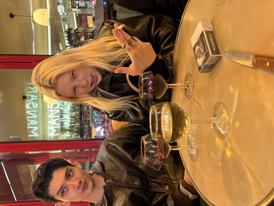A young woman with blonde hair and a black leather jacket holds a cigarette and winks. She sits with a young man wearing a similar jacket at a round table. The table holds a deck of cigarettes, a knife, and three wine glasses filled with white and red wine. The setting appears to be a restaurant, evidenced by the blurred backdrop of shelves stocked with bottles. A neon sign is visible in the window.