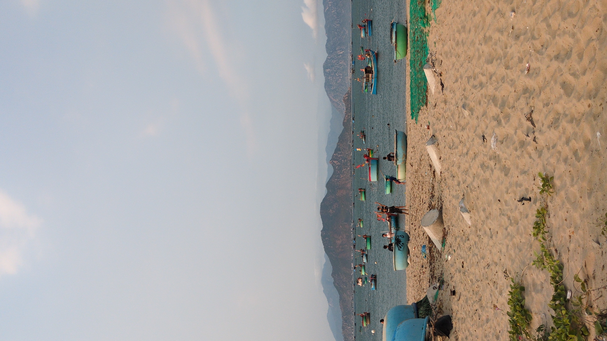 The image depicts a coastal scene at Phan Rang – Tháp Chàm, Vietnam. Several small fishing boats, painted in shades of blue and green, are docked along the sandy beach. Some people are near the boats, likely fishermen or locals. In the background, a mountainous landscape stretches along the coastline. The foreground shows the sandy beach with scattered debris and vegetation. The sky is pale blue with faint clouds.