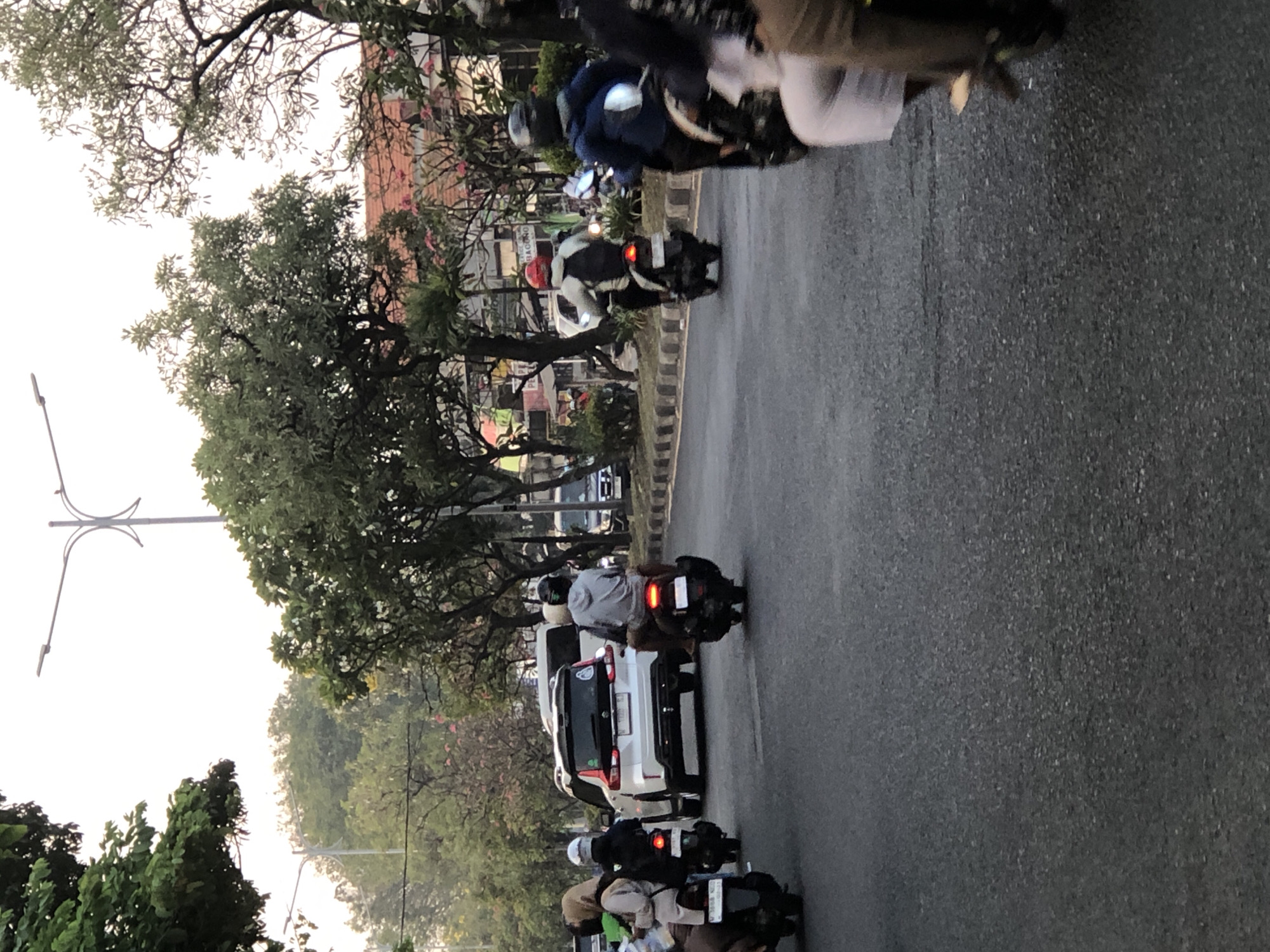 This image depicts a street scene in Sumorame, Indonesia, likely during the daytime with overcast skies. The foreground features an asphalt road with several motorcycles and their riders. On the left, a motorcycle carries two individuals, one wearing a light-colored hijab and the other a dark jacket and helmet. Further down the road, another motorcycle has a single rider in a light gray hoodie. In the right foreground, parts of another motorcycle and two individuals are visible, one wearing a light-colored skirt and the other a dark jacket.
In the middle ground, a white SUV is driving in the same direction as the motorcycles. To the right of the SUV, a person on a motorcycle wearing a red helmet and a light-colored jacket is visible. Further in the background, more vehicles and buildings can be seen. The environment includes trees with lush green foliage lining the sides of the road, and streetlights are present above the traffic. The overall impression is of a moderately busy street with typical urban traffic.