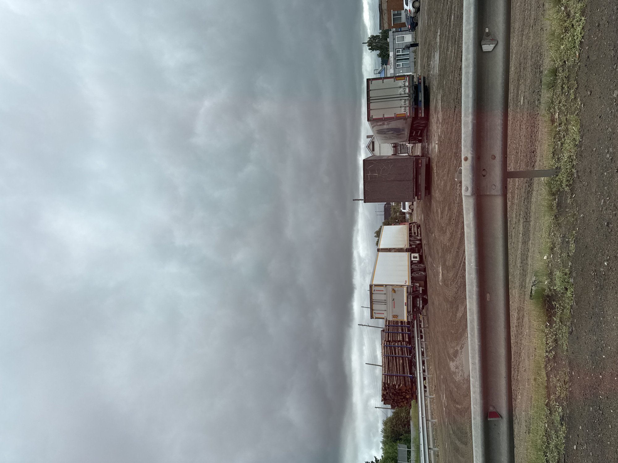 This is a ground-level, eye-level, vertical, wide shot of a logging area and truck yard under a cloudy sky. In the foreground, a metal guardrail runs horizontally across the bottom of the frame, separating the viewer from a muddy, dirt area with patches of grass. Behind the guardrail, several large trucks are parked. The leftmost truck is a logging truck, carrying a large load of cut logs secured with blue straps. Next to it is a white semi-trailer truck. Further to the right are two more semi-trailer trucks, one a dark color and the other white. In the background, there is a small building with "ПРОДУКТЫ" (Products) signage, a few parked cars, and trees. The ground is wet and appears to be a mix of mud and dirt, with tire tracks visible. The sky is overcast with heavy, dark clouds. The scene suggests a commercial or industrial area, likely a log processing or distribution center, in Alnashi, Russia. There are no people actively engaged in actions.