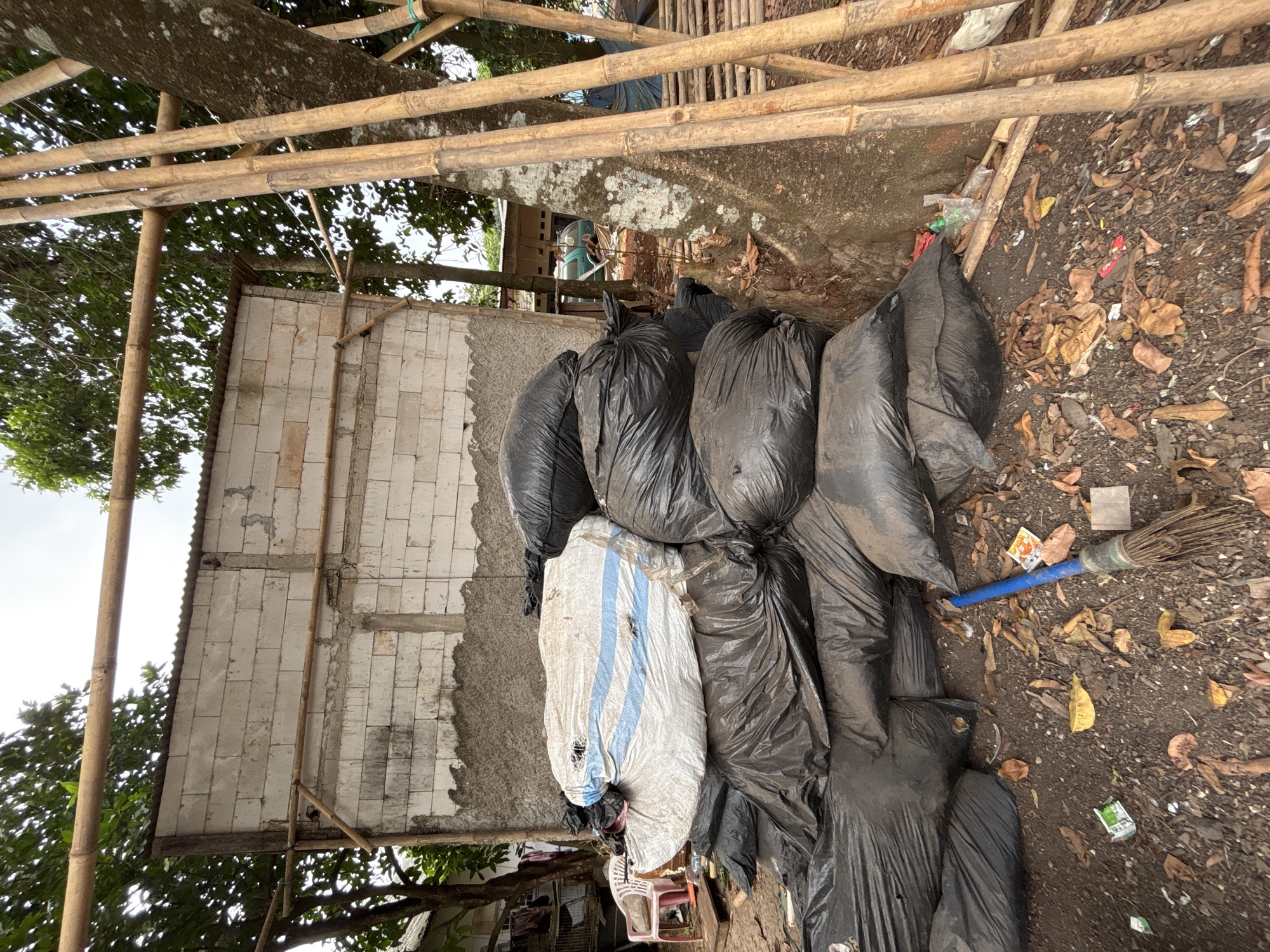 The image shows an outdoor scene at a construction site in Taman Rahayu, Indonesia, with a pile of black trash bags and one white and blue striped bag stacked in front of a partially constructed building. The building appears to be made of white cinder blocks with a corrugated metal roof. Bamboo poles are visible in the foreground and background, supporting the roof structure and leaning against a large tree. The ground is covered with dirt, dry leaves, and scattered debris, including a blue-handled broom and some small pieces of trash. A pink plastic chair is visible to the left of the pile of bags. The overall scene depicts a messy and unkempt area, likely a worksite or storage area for waste materials.