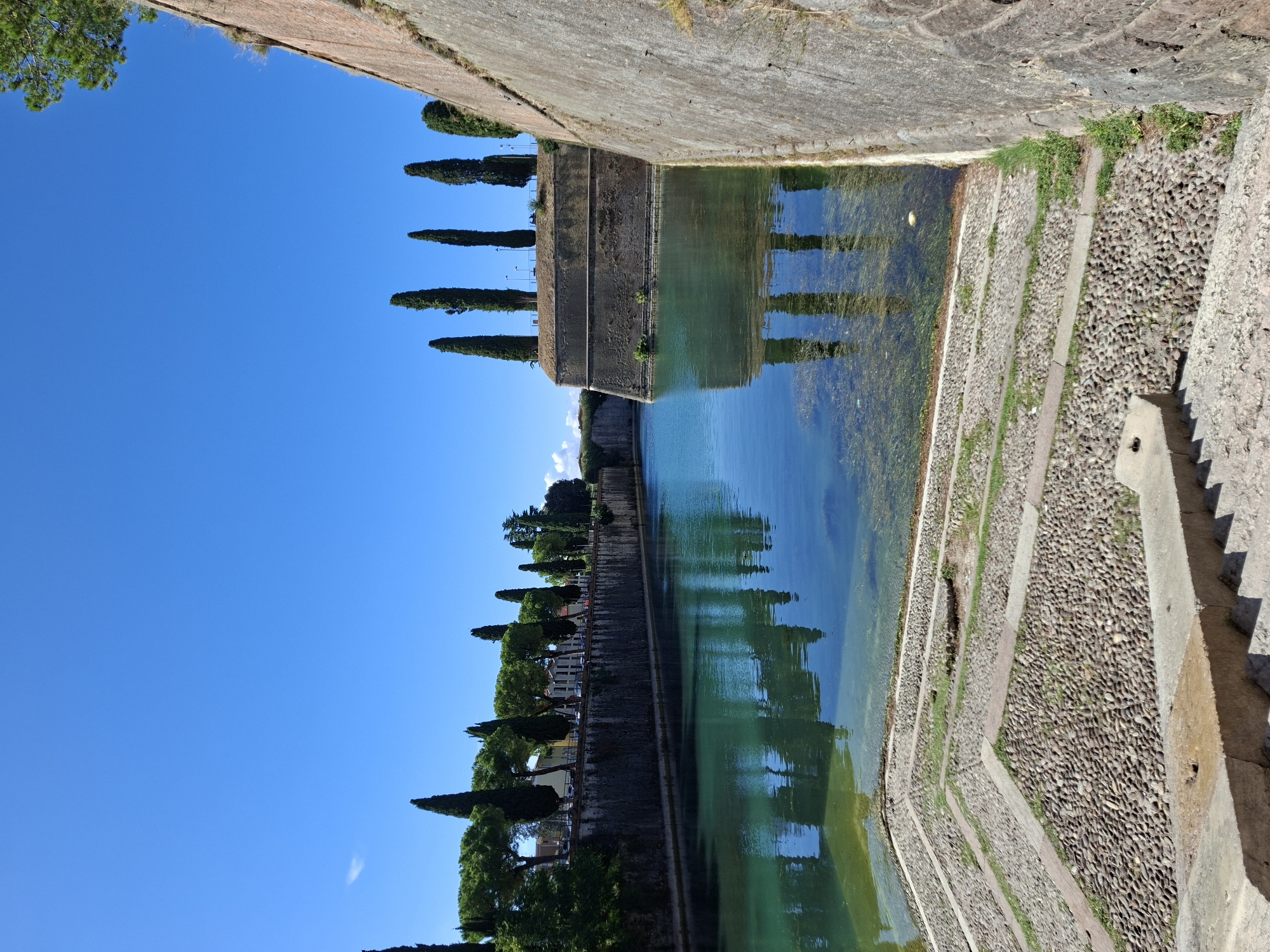 The image shows a view of the moat surrounding the historic fortress of Peschiera del Garda, Italy. The water in the moat is clear and turquoise, with visible aquatic plants and a yellow object floating on the surface. The moat walls are constructed of rough-hewn stone, and in the foreground, there are stone steps leading down to the water, bordered by paths of small, rounded pebbles interspersed with grass.
Along the opposite bank of the moat, a stone wall lines the perimeter, topped with a row of tall, slender cypress trees and a variety of other trees and greenery. Beyond the trees, buildings with light-colored facades are visible, with parked cars suggesting an urban setting.
The sky is a vibrant, clear blue, indicating a sunny day. The reflections of the trees and the fortress wall are clearly visible in the still water of the moat. The overall scene depicts a well-preserved historical site with natural beauty.