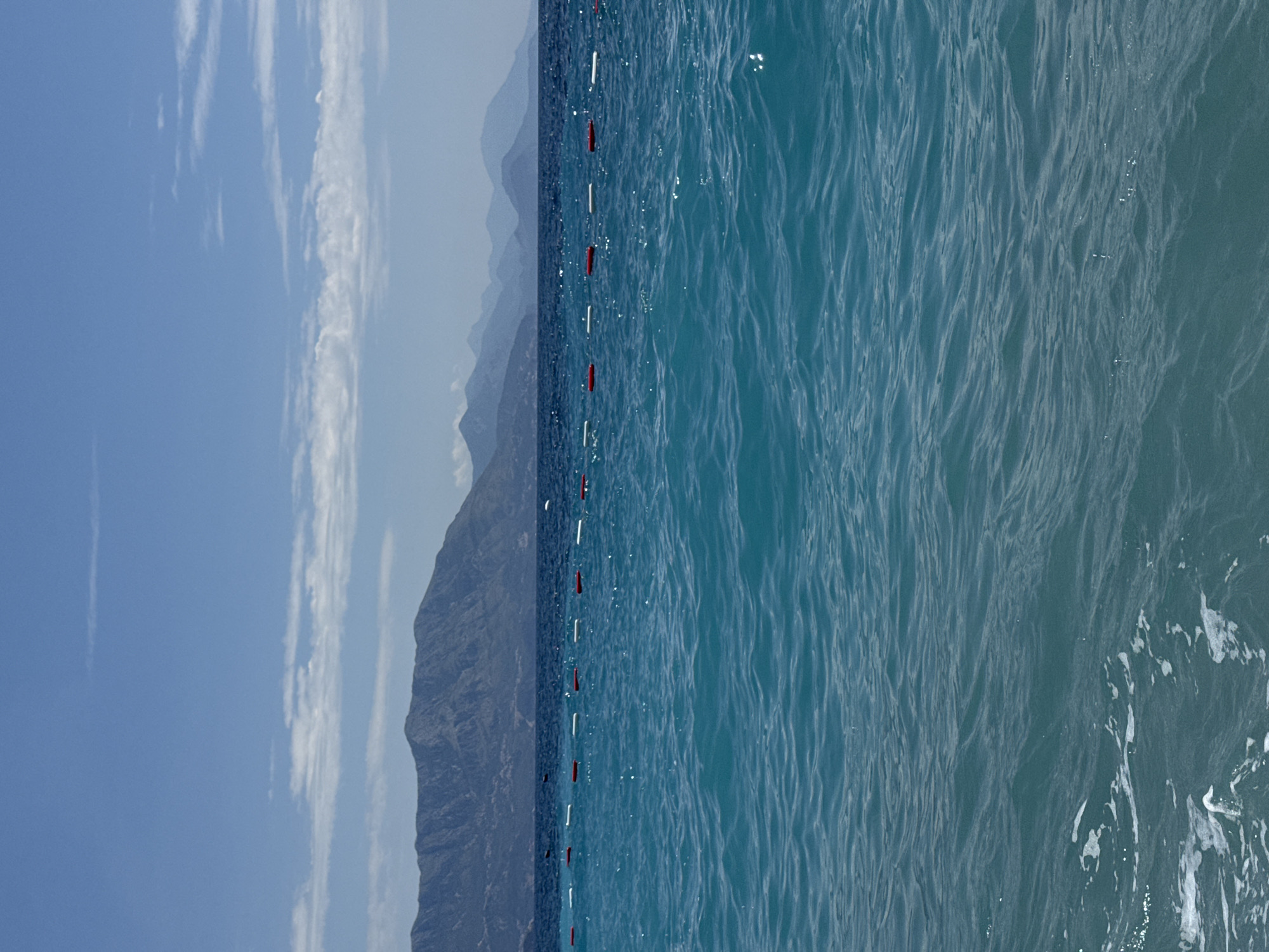 The image depicts a serene coastal scene with a clear blue sky dotted with scattered white clouds. In the foreground, the turquoise water of the Adriatic Sea is gently rippling, with some white foam visible along the bottom edge. A line of buoys, alternating between red and white, stretches horizontally across the middle ground, marking a boundary in the water. Beyond the buoys, the sea deepens to a darker blue. In the background, a chain of imposing mountains rises, their slopes covered in lush greenery, with the farthest peaks appearing hazy in the distance. The overall impression is one of natural beauty and tranquility. This scene is located in Prijevor, Montenegro.