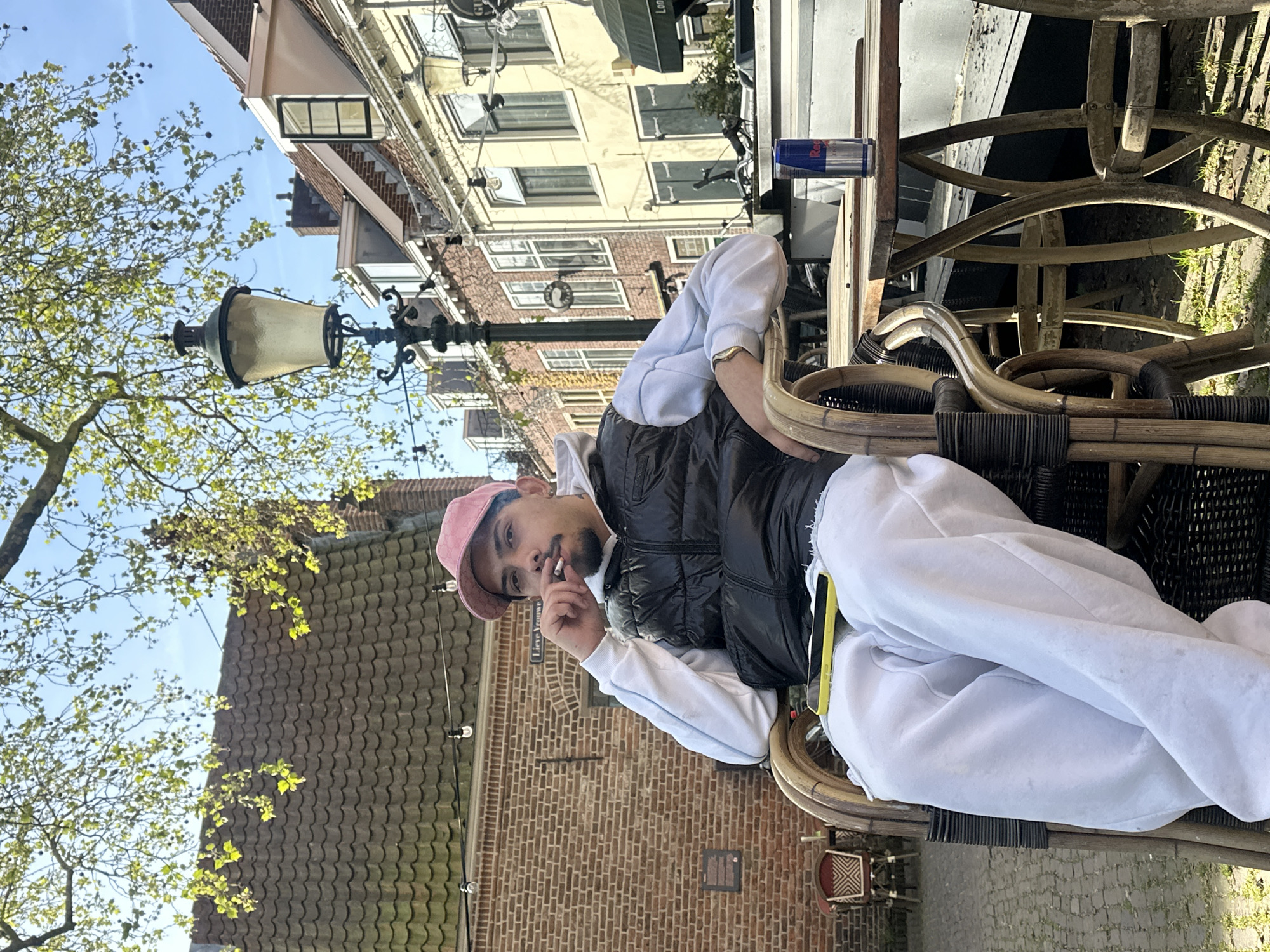 The image depicts a young man sitting at an outdoor cafe table on a sunny day in Amersfoort, Netherlands. The setting features traditional brick buildings with distinct architectural styles, a clear blue sky, and a tree with fresh green leaves, suggesting spring or early summer.
The main subject is a young man with a mustache and beard, wearing a pink baseball cap, a white hoodie, and a black puffer vest. He is seated in a rattan-style chair, holding a lit cigarette to his mouth and looking directly at the viewer with a neutral expression. On the wooden table in front of him, a blue and silver Red Bull can is visible.
Notable objects in the background include an ornate black lamppost with a large, textured glass light fixture, and several multi-story buildings with brick facades and numerous windows. A street sign on a brick wall behind the man is partially visible, displaying the text "Lieve Vrouwe," which indicates the location. The ground, partially visible through the chairs, appears to be paved with cobblestones or similar patterned stones. The bright light indicates it is daytime, under clear weather conditions.