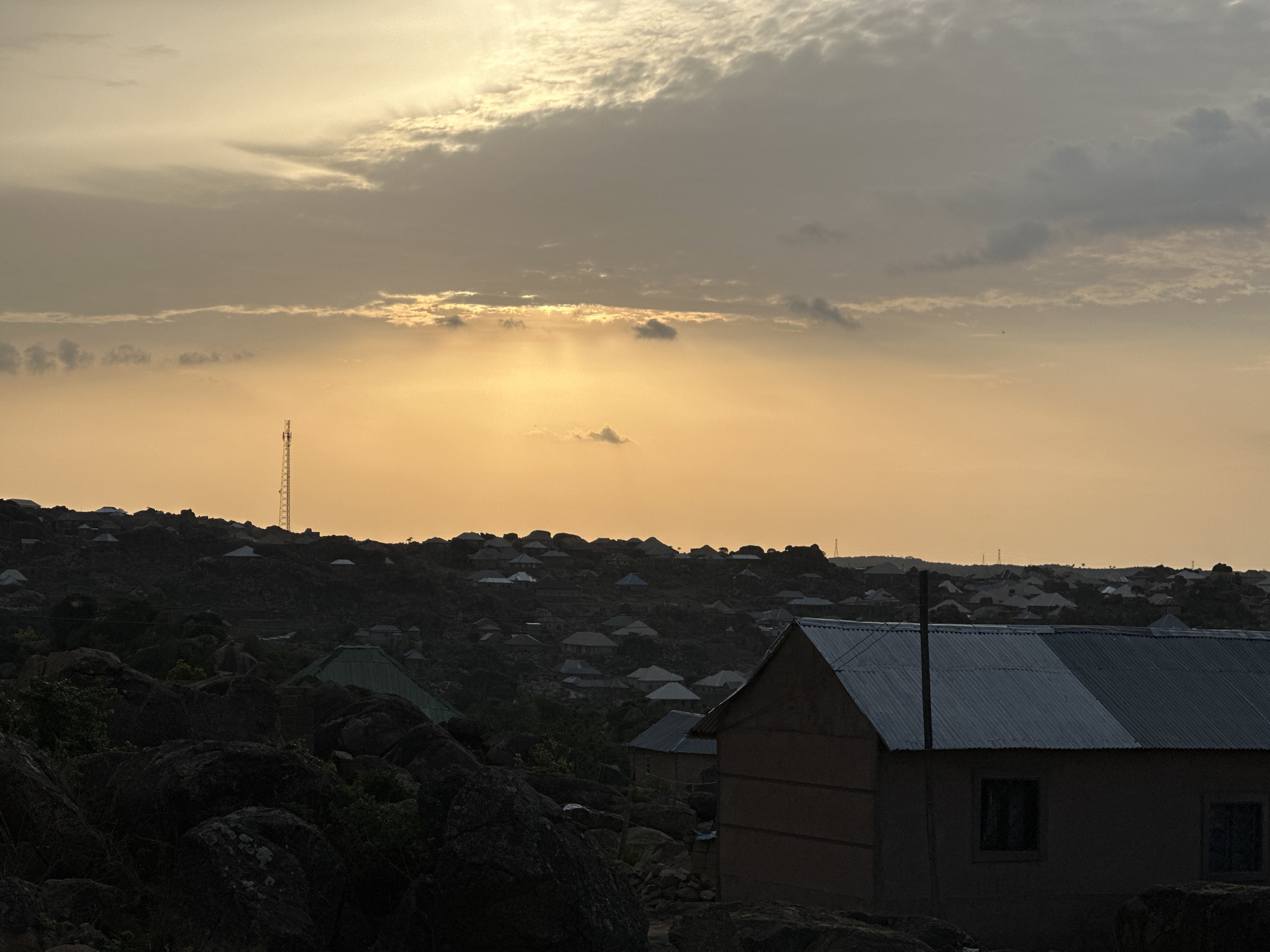 The image depicts a landscape in Jos, Nigeria at dusk. In the foreground, there are large rocks and a building with a corrugated metal roof and a simple, likely earthen construction. In the midground, numerous smaller buildings and houses are scattered across a hilly terrain, appearing densely populated. A telecommunications tower is visible on one of the hills. The sky is filled with clouds, and the sun's light is partially obscured, creating an orange/yellow glow over the scene. The lighting suggests the end of the day. No people are directly visible in the image.