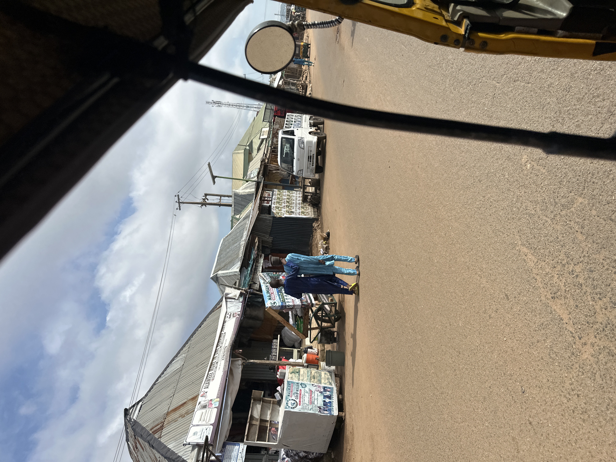 The image depicts a street scene in Jos, Nigeria, as viewed from the perspective of a passenger in a yellow auto-rickshaw. In the foreground, the right side of the auto-rickshaw's frame and a portion of its yellow body are visible. A dark metal bar, likely part of the vehicle's structure, runs vertically through the left side of the frame.
The street itself is paved with asphalt and appears to be a commercial area. On the left side of the street, several market stalls or small shops line the road. These structures have corrugated metal roofs and appear to be made of various materials. One prominent stall features a large white sign with text and images, including what appears to be information about "NAJMAH COLD KILLER." Adjacent to this stall is a white cart with shelves.
Two men are walking away from the camera on the left side of the street, dressed in traditional African attire. One is wearing a dark blue robe and a dark cap, while the other is in a light blue robe and a similar cap. They are walking on the dirt shoulder next to the paved road.
Further down the street on the left, a white commercial truck with a flatbed is parked. More market stalls and shops are visible along the right side of the street. In the distance, a person is walking on the right side of the road. The sky above is partly cloudy with patches of blue visible. The overall environment suggests a bustling marketplace or a commercial district within the city of Jos.