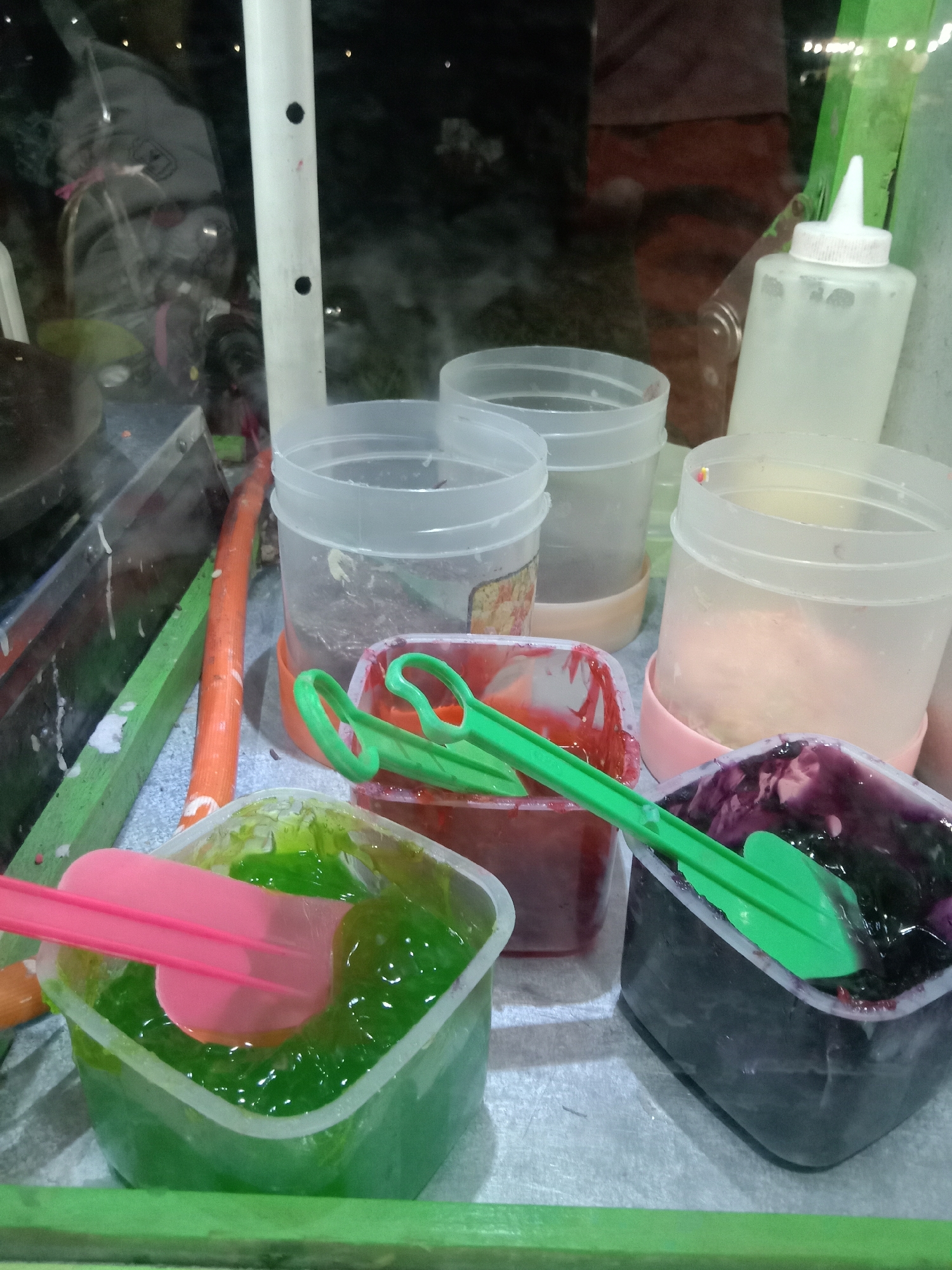 The image depicts a close-up view of a food stall display, likely in Stiaji, Indonesia. In the foreground, several transparent plastic containers hold colorful liquids. The container on the far left contains a bright green viscous substance, with a pink spatula submerged in it. To its right is another container with a red liquid, and further to the right, a container filled with a dark purple substance, possibly a fruit puree or jam, with a green utensil resting on top. Behind these, three tall, clear plastic cylinders, some with lids, are arranged, appearing to hold more ingredients or display items. One of these cylinders contains a light pink, possibly powdered, substance. To the right of these, a white squeeze bottle with a red-capped lid is visible. The overall setting suggests a preparation area for street food, possibly crepes or pancakes, given the presence of spatulas and liquid ingredients. The background is dark and out of focus, but hints at reflections of lights and possibly people or equipment within the stall.