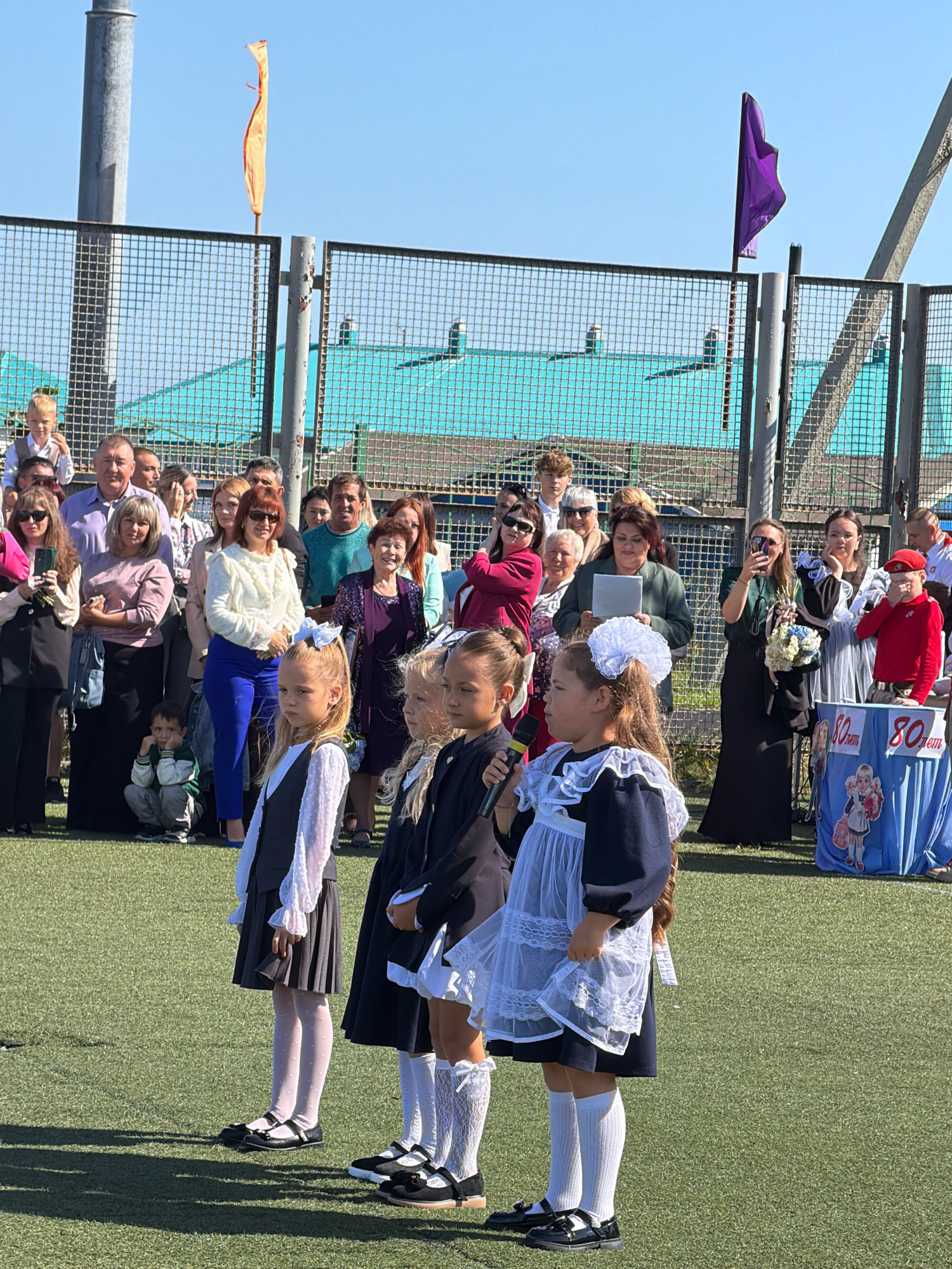 The image depicts an outdoor event on a sunny day, likely a school assembly or celebration, on a green turf field. A group of people, predominantly adults and children, are gathered. Several young girls in school uniforms, including navy dresses with white collars and aprons, and some in white blouses and dark skirts, are positioned in the foreground. One girl is speaking into a microphone, while others stand attentively. Adults are standing behind the children, some observing the event, others holding phones, and one holding a tablet. A blue table decorated with "80 лет" (80 years) is visible on the right, adorned with a picture of a girl with a backpack. In the background, a chain-link fence separates the field from a building with a teal roof and a light-colored structure behind it. Two flags, one orange and one purple, are hoisted on poles against a clear blue sky. The general atmosphere suggests a community gathering or a formal school occasion. The location is identified as Yuzhno-Kurilsk, Russia.