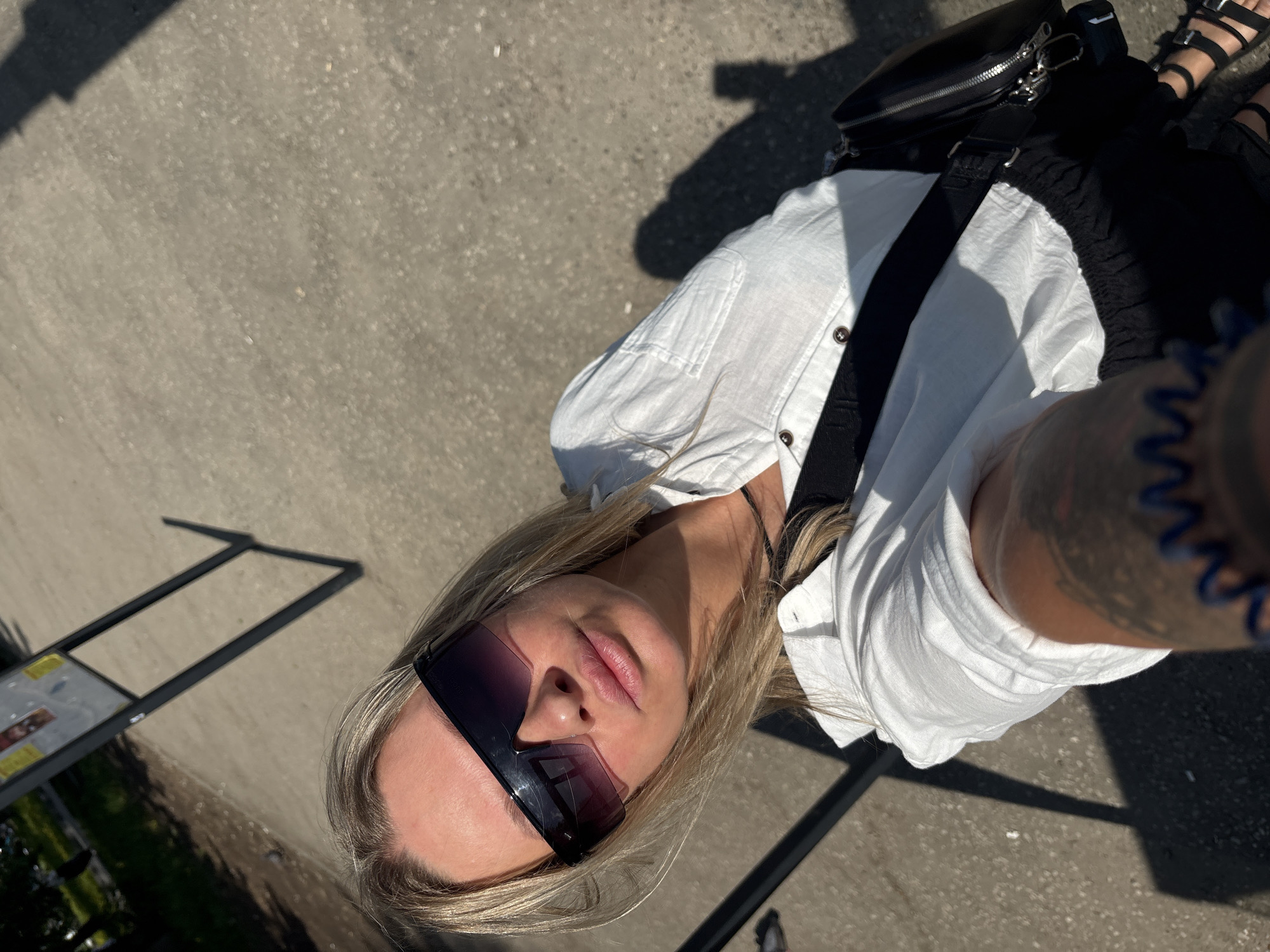 A young woman is taking a selfie outdoors, likely in a public area. She is wearing sunglasses, a white button-down shirt, and a black bag. Her blonde hair is loose around her face. The background appears to be a concrete surface with some metal structures, possibly parts of street furniture or signage. Although the precise location within Perm, Russia cannot be determined from this single image, the presence of street furniture and signs suggests it could be a park, square, or pedestrian zone.