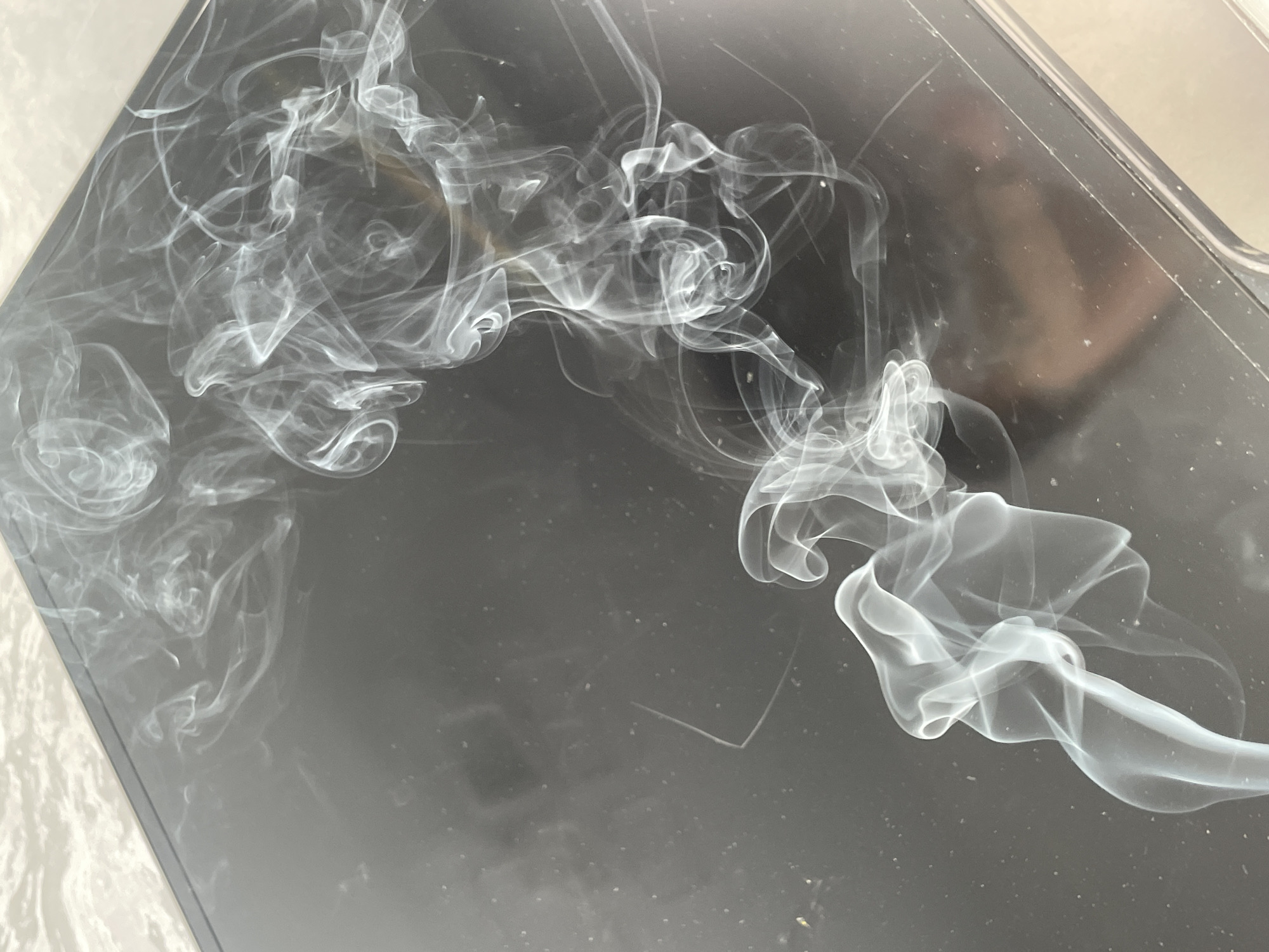 The scene depicts white-grey smoke rising and swirling above a dark, reflective surface, likely a table or electronic device. This surface exhibits numerous small dust particles and faint linear scratches. In the upper-left corner, a lighter, textured, off-white surface, possibly a wall or countertop, abuts the dark surface along a diagonal line. The smoke plumes vary in density and form, creating intricate patterns as they disperse. A faint, blurred reflection of a human head and shoulders is visible in the lower-right quadrant of the dark surface, indicating the presence of an individual. The specific address or building within Izhevsk, Russia, where this interior scene is located is not identifiable from the visual data.