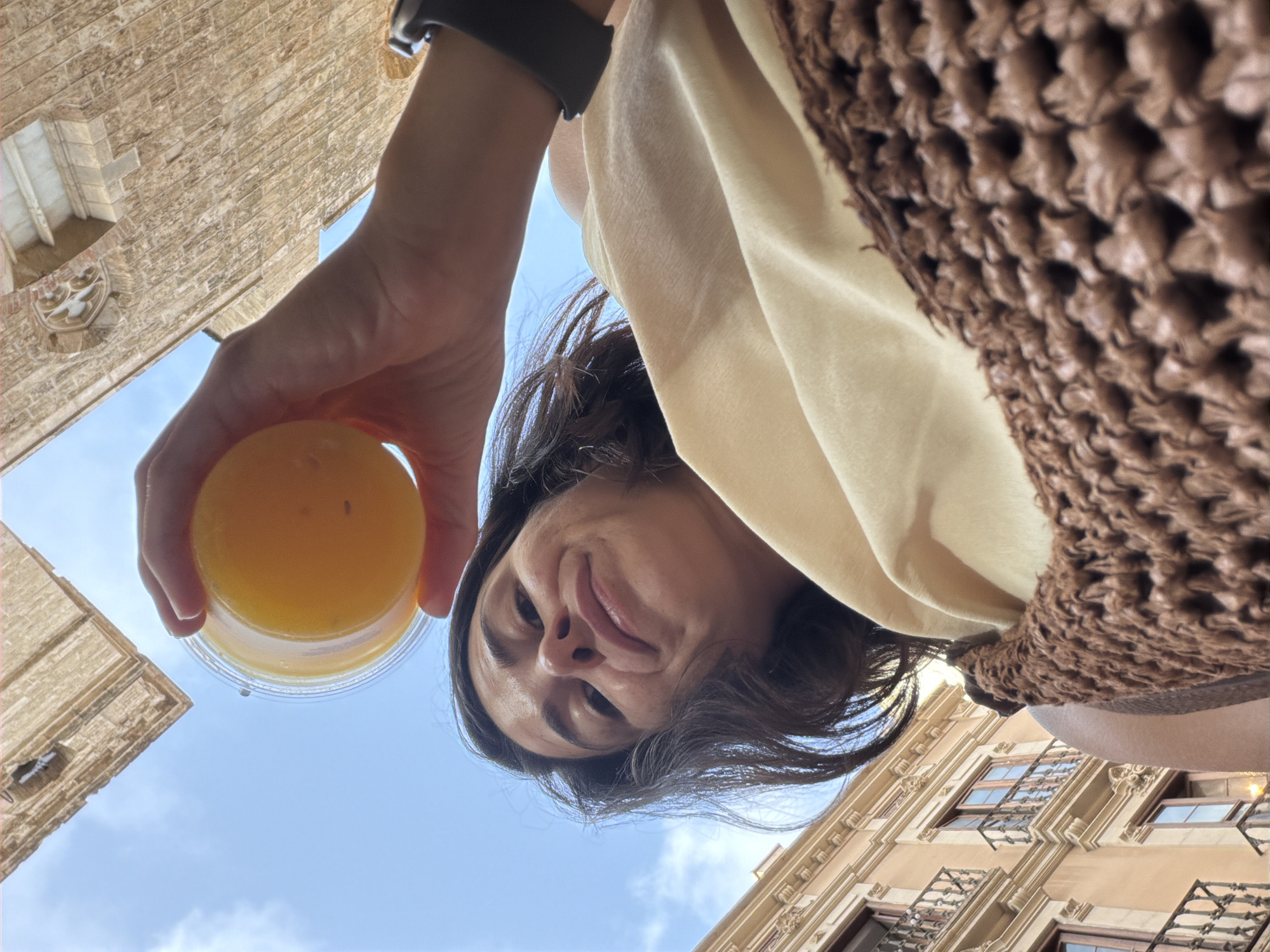 In an outdoor, low-angle shot, a woman is holding up a clear plastic cup filled with orange juice. She is wearing a tan top and a brown woven bag rests on her shoulder. A dark smartwatch is visible on her left wrist. The background features the sky with scattered clouds and the facade of an old building with balconies. The overall lighting suggests it is daytime.