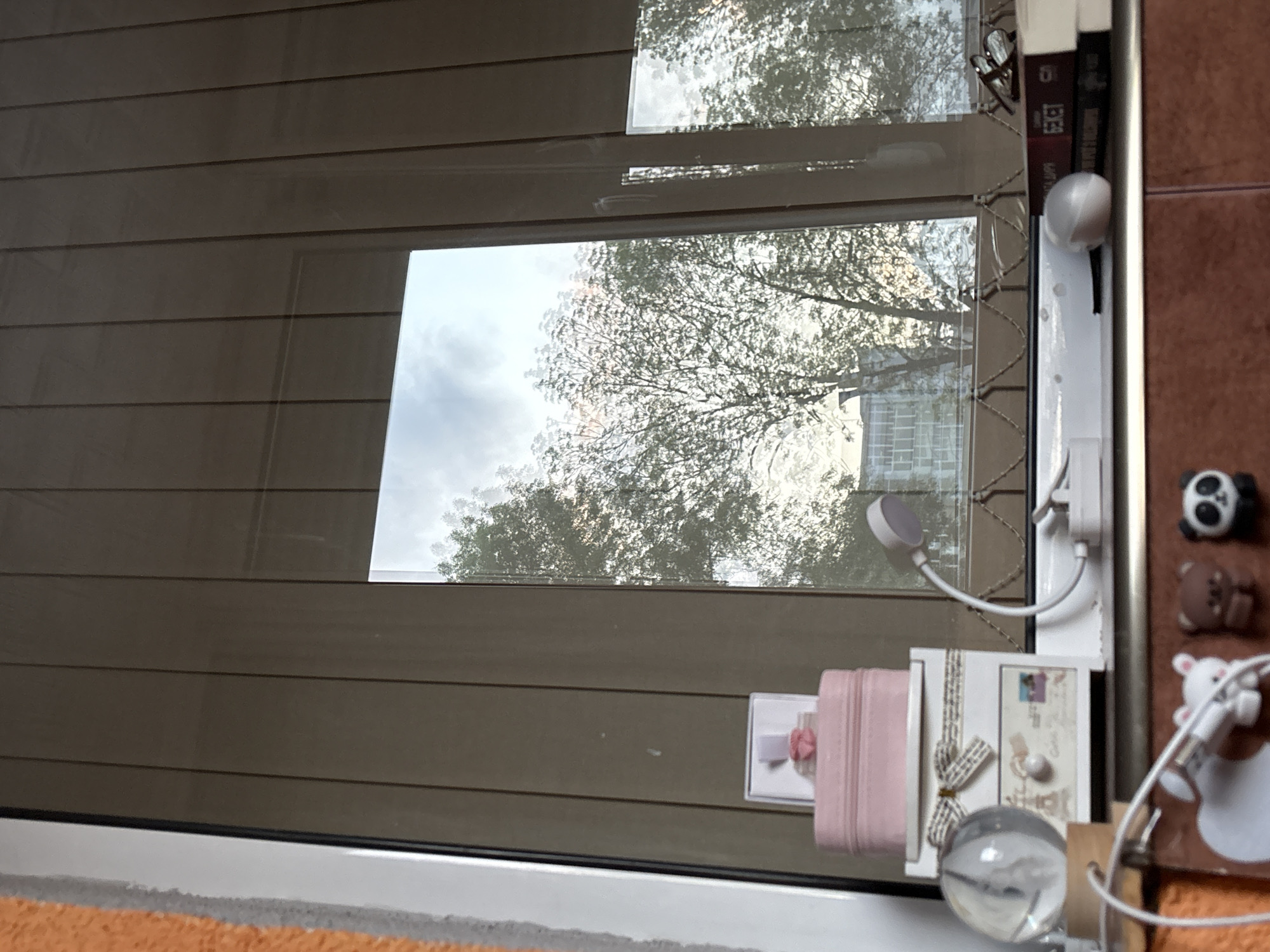 The image shows a view through a window with closed blinds. Through the window, trees are visible, suggesting an outdoor environment. The window is adorned with various items, including decorative boxes, a postcard, a clear glass sphere, a charging cable and other small trinkets. The overall setting gives the impression of a residential space with personalized decorations. It is not possible to determine if the scene is located in Panagyurishte, Bulgaria, or anywhere else from the image alone. There are no people visible.