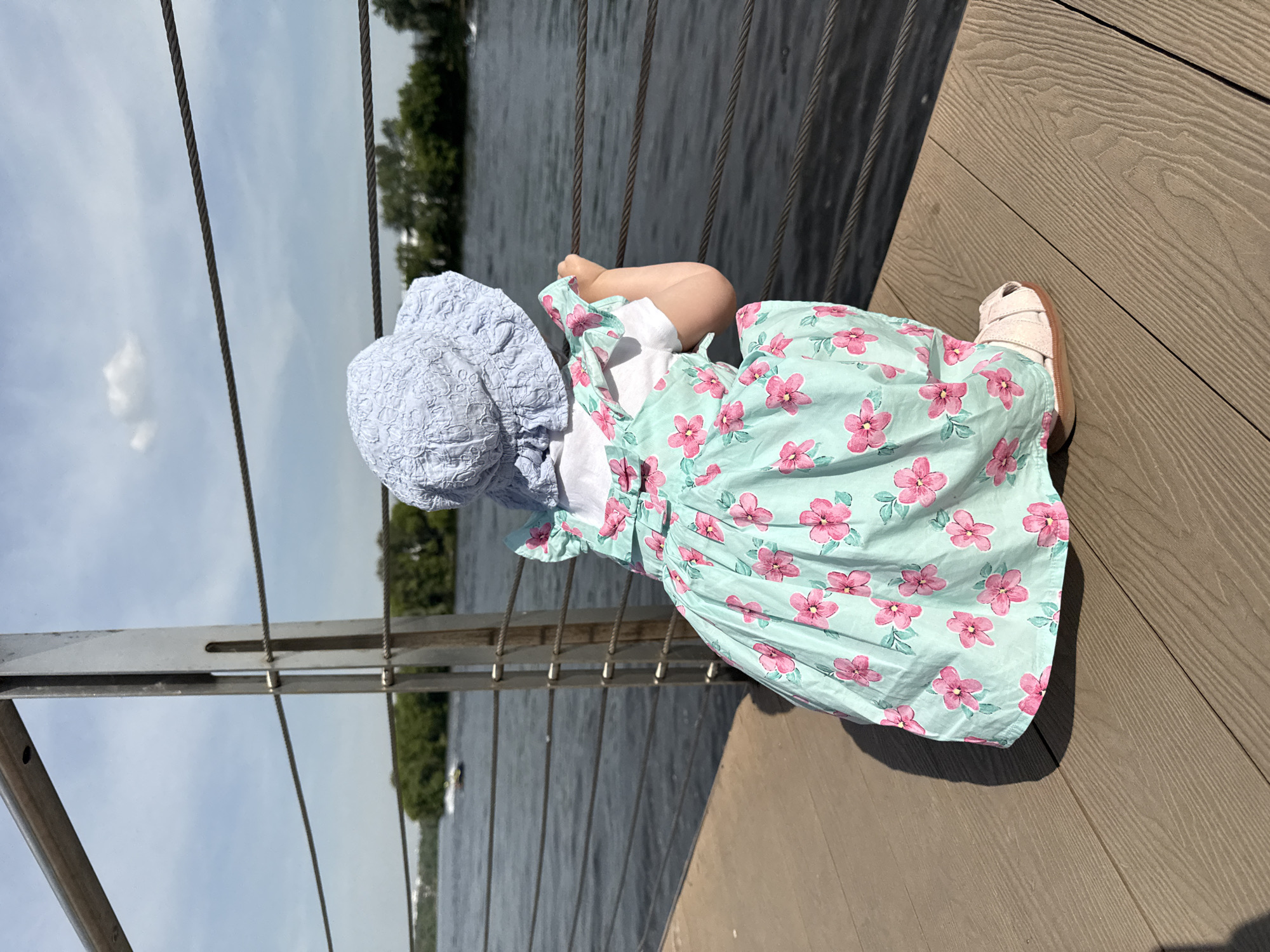The image depicts a young child, seen from behind, squatting on a light brown composite wood deck while looking out over a wide river. The child wears a light blue, textured bucket hat, a white short-sleeved top, and a mint green sundress patterned with scattered pink flowers and green leaves. The dress features ruffled shoulder straps and a small bow detail on the upper back. Light-colored, closed-toe sandals are visible on the child's feet.
The child's hands are gripping the lowest of three visible dark metal cables that form a railing. These cables are stretched between sturdy, brushed metal vertical posts. Beyond the railing, the dark, rippled surface of a wide river extends into the distance. A small, light-colored motorboat or jet ski is visible on the far left side of the river, leaving a white wake. The opposite bank is lined with dense green trees and foliage, with hints of distant buildings behind them. The sky is a light blue with scattered white clouds. Strong sunlight is indicated by the prominent shadow of the child cast on the deck. The scene is consistent with a modern pedestrian embankment along the Moskva River in Moscow, Russia.