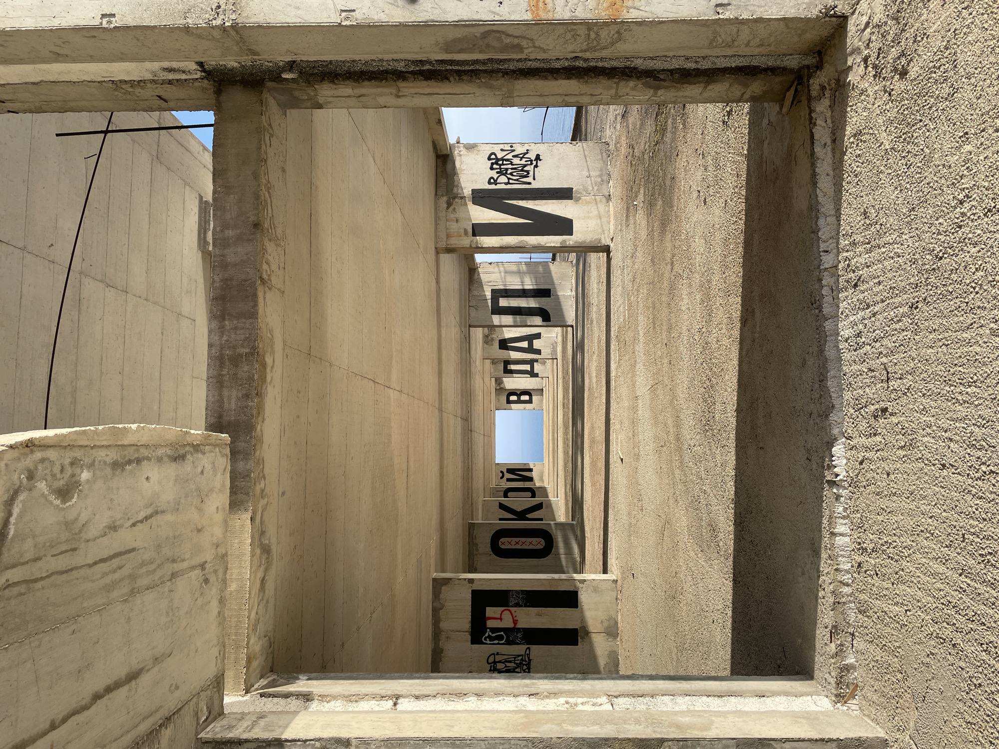 The image depicts a partially constructed or dilapidated concrete structure, likely a building, in Perazića Do, Montenegro. The view is framed by an incomplete doorway or window, constructed of weathered concrete. The perspective looks through a series of concrete beams or supports, spaced apart to create a corridor effect. Each beam is marked with Cyrillic letters, stacked vertically. Some beams feature additional graffiti. Light streams through the gaps between the beams, revealing a glimpse of the sky. The overall scene conveys a sense of abandonment and decay, set against the backdrop of an unfinished or ruined architectural project.