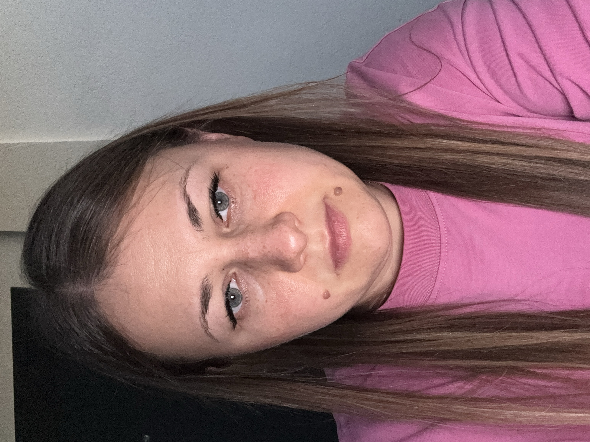 A woman with medium-length brown hair and fair skin is shown from the shoulders up, facing the camera directly. She is wearing a bright pink crewneck sweatshirt. Her eyes are light blue and her eyebrows are dark. She has prominent eyelashes and light makeup. There are two moles visible on her face, one on her left cheek and one on her right cheekbone. The background is split between a dark vertical surface on the left and a light gray, textured wall on the right.