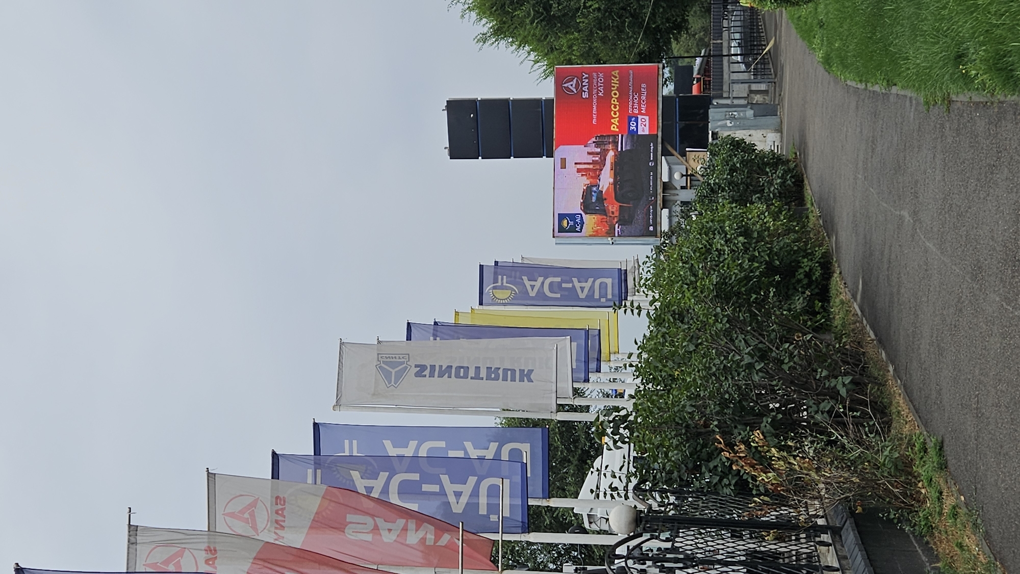 A digital billboard displays an advertisement for SANY pneumohydraulic excavators. The advertisement highlights a 30% down payment and a 20-month payment plan. The billboard is situated in Almaty, Kazakhstan, likely in a commercial area given the presence of multiple large flags with branding. The flags, in red, blue, white, and yellow, are displayed in a row, suggesting a dealership or exhibition. The environment is an outdoor setting with a paved walkway, green bushes, and trees. The sky is overcast.