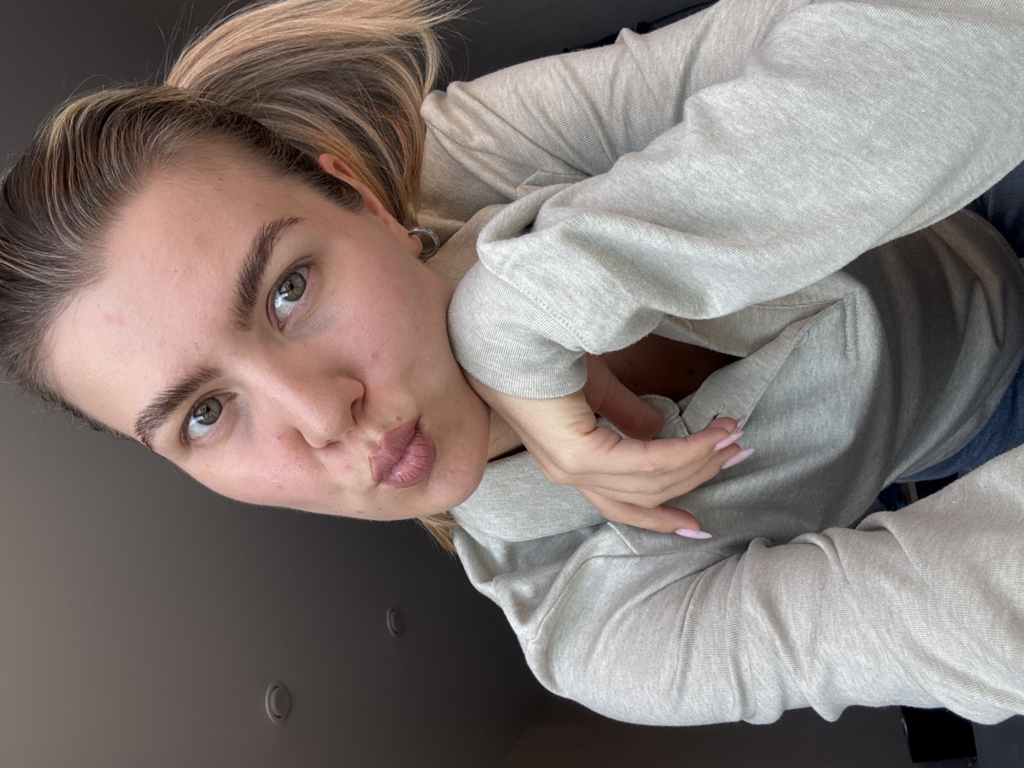 A casual selfie features a light-skinned individual, likely a young woman, from an upward-angled perspective. Her light brown hair is pulled back in a ponytail, revealing her left ear with a small, circular earring. She has expressive green eyes and dark eyebrows. Her lips are puckered in a "kissing" or "duck face" pose. She is wearing a light gray, textured long-sleeved top, possibly a hoodie or sweatshirt, with her left hand resting gently near her chin. Her fingernails are long and painted a light pink. The background is a plain, dark gray or black surface, possibly a wall or furniture, with a few subtle, circular indentations visible. The lighting is soft and even, suggesting an indoor environment. The overall mood is casual and playful. There is no visible text.