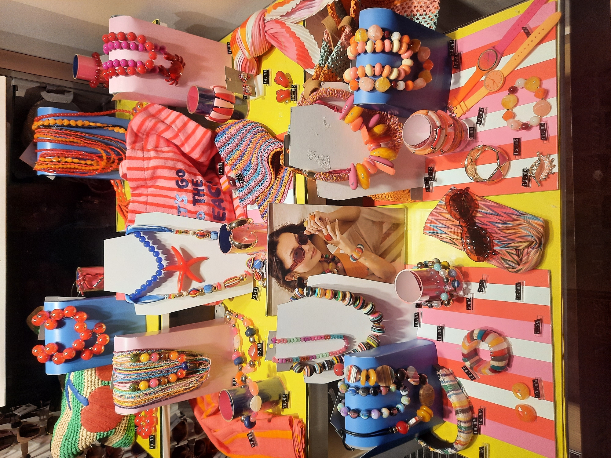 The image displays a brightly colored jewelry and accessory store window in Rosenheim, Germany. The display features an assortment of necklaces, bracelets, earrings, sunglasses, and a tote bag, all with a vibrant, summery aesthetic. The items are arranged on pink, blue, and yellow pedestals and a striped surface, creating a cheerful and eye-catching presentation. Several price tags are visible throughout the display. The overall impression is one of a boutique offering trendy, statement accessories.