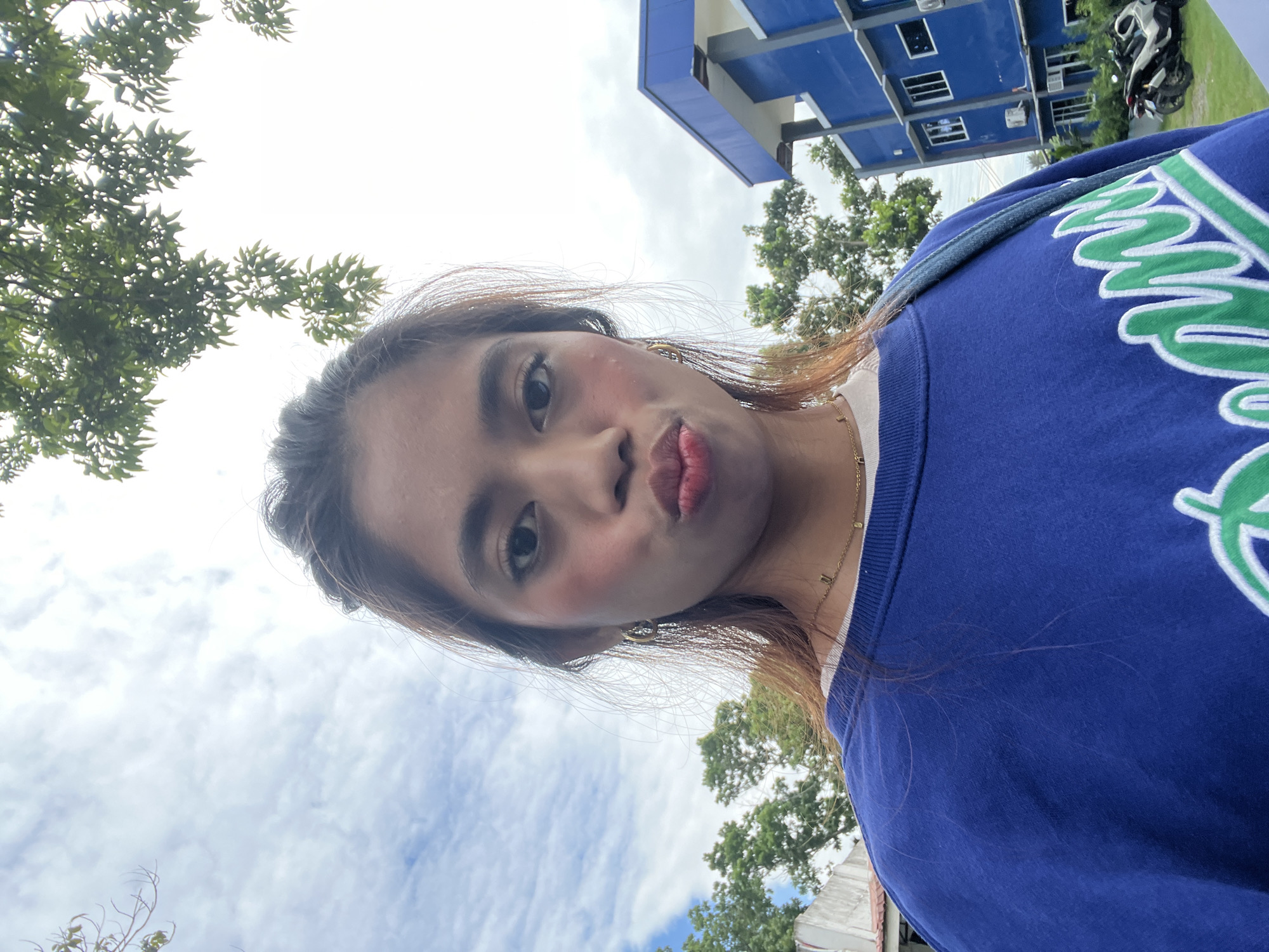 A young woman with brown hair is taking a selfie outdoors. She is wearing a blue sweatshirt and a necklace. Her lips are pursed as if she is blowing a kiss. In the background, there is a blue building and trees. The sky is cloudy. The location is identified as Panabo City, Philippines.