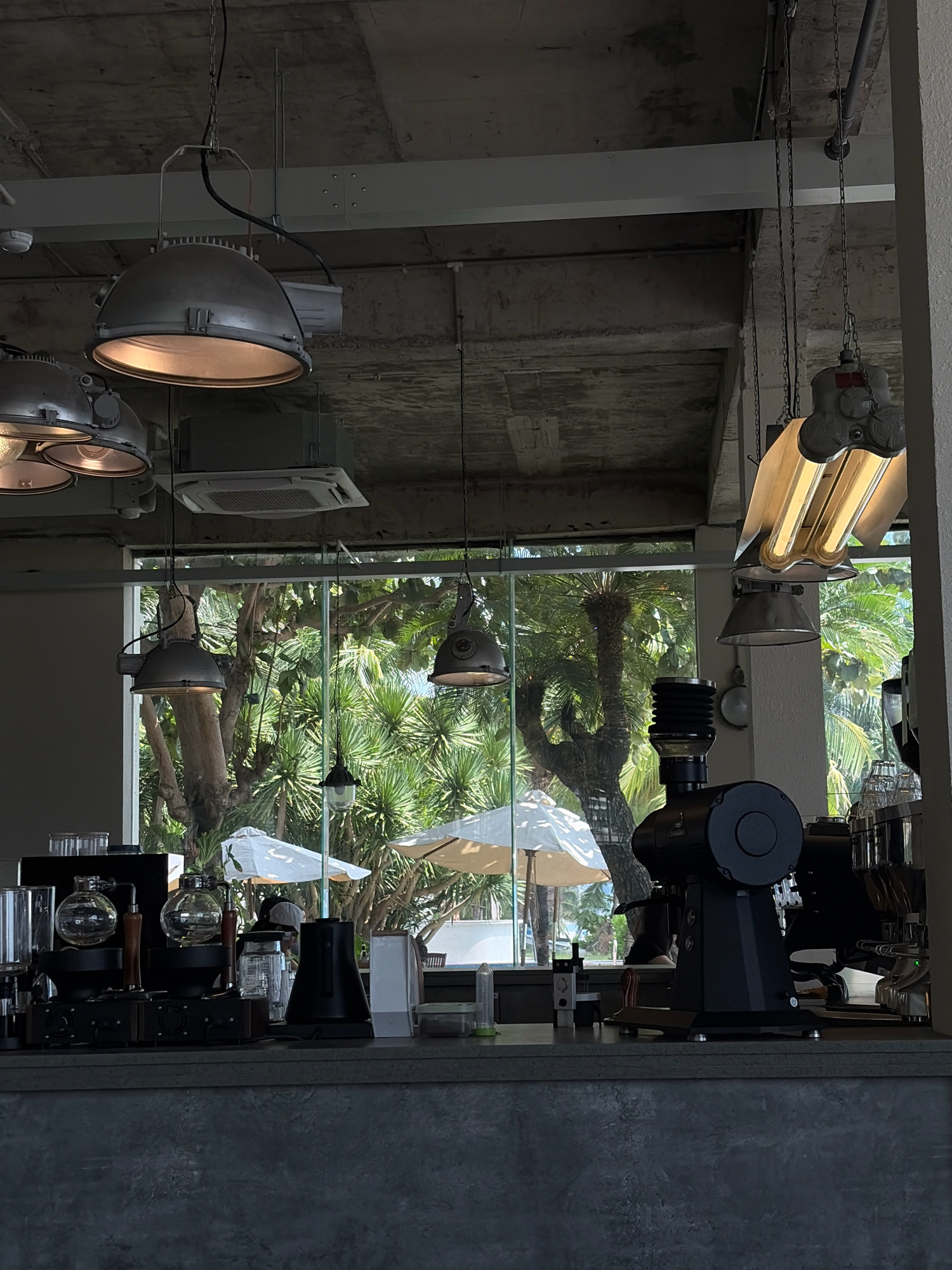 The image shows the interior of a modern cafe, possibly located in the city of Đà Nẵng, Vietnam, given the tropical foliage visible through the large windows. The cafe features an industrial aesthetic with exposed concrete ceilings and industrial-style pendant lighting. A grey concrete counter dominates the foreground, displaying various coffee-making equipment, including a large black coffee grinder, siphon coffee makers, and glassware. In the background, through the windows, lush green trees and white umbrellas suggest an outdoor seating area, possibly near a beach. The overall atmosphere appears relaxed and contemporary. There are no people clearly visible in the frame.