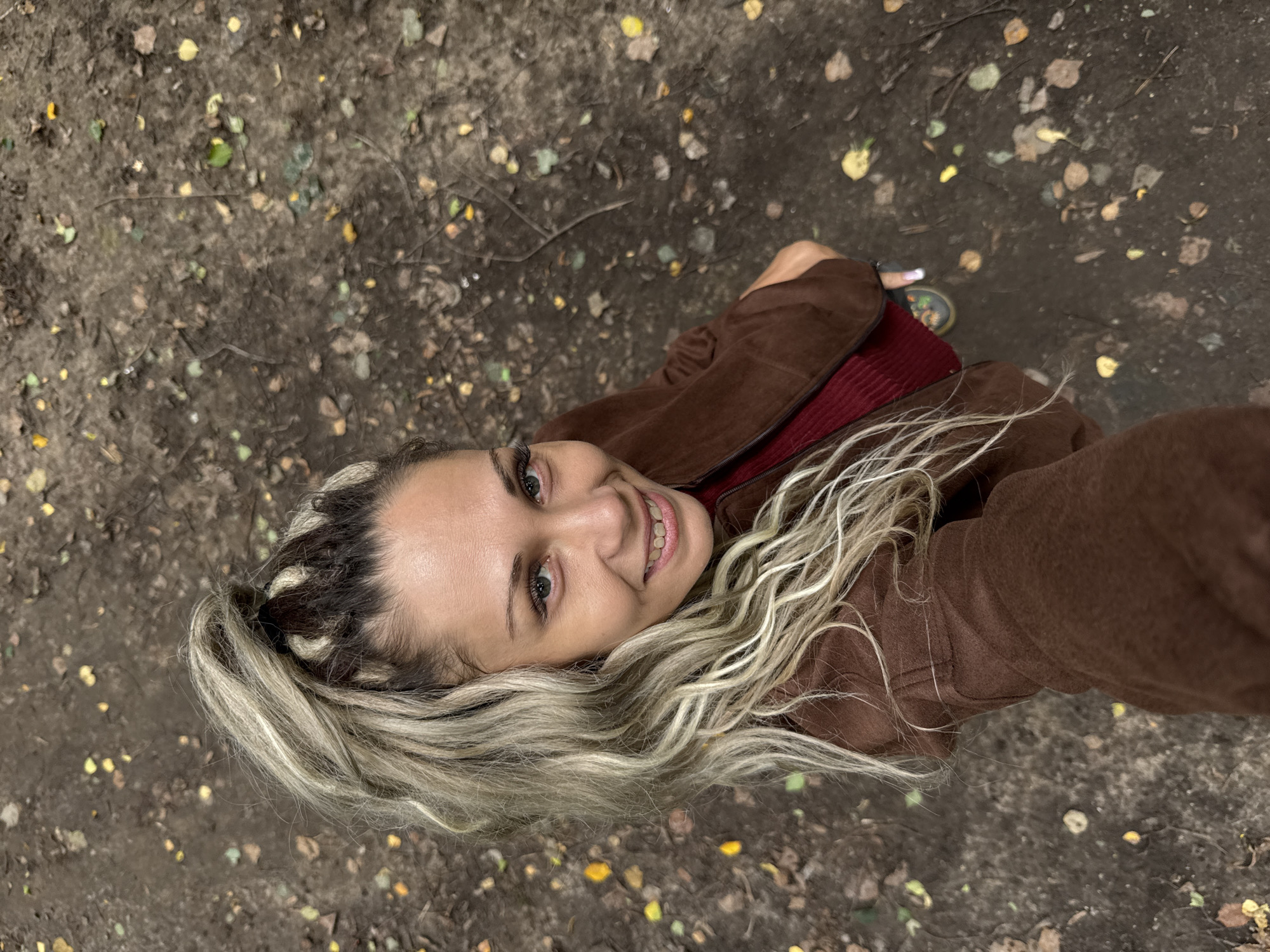 A woman with blonde dreadlocks pulled into a high ponytail is taking a selfie outdoors on a dirt path. She is wearing a brown jacket over a red top. The ground is covered with dirt, fallen leaves, and some small yellow and green leaves. The woman is smiling and looking at the camera. The location is the city of Rusanovo, Russia.