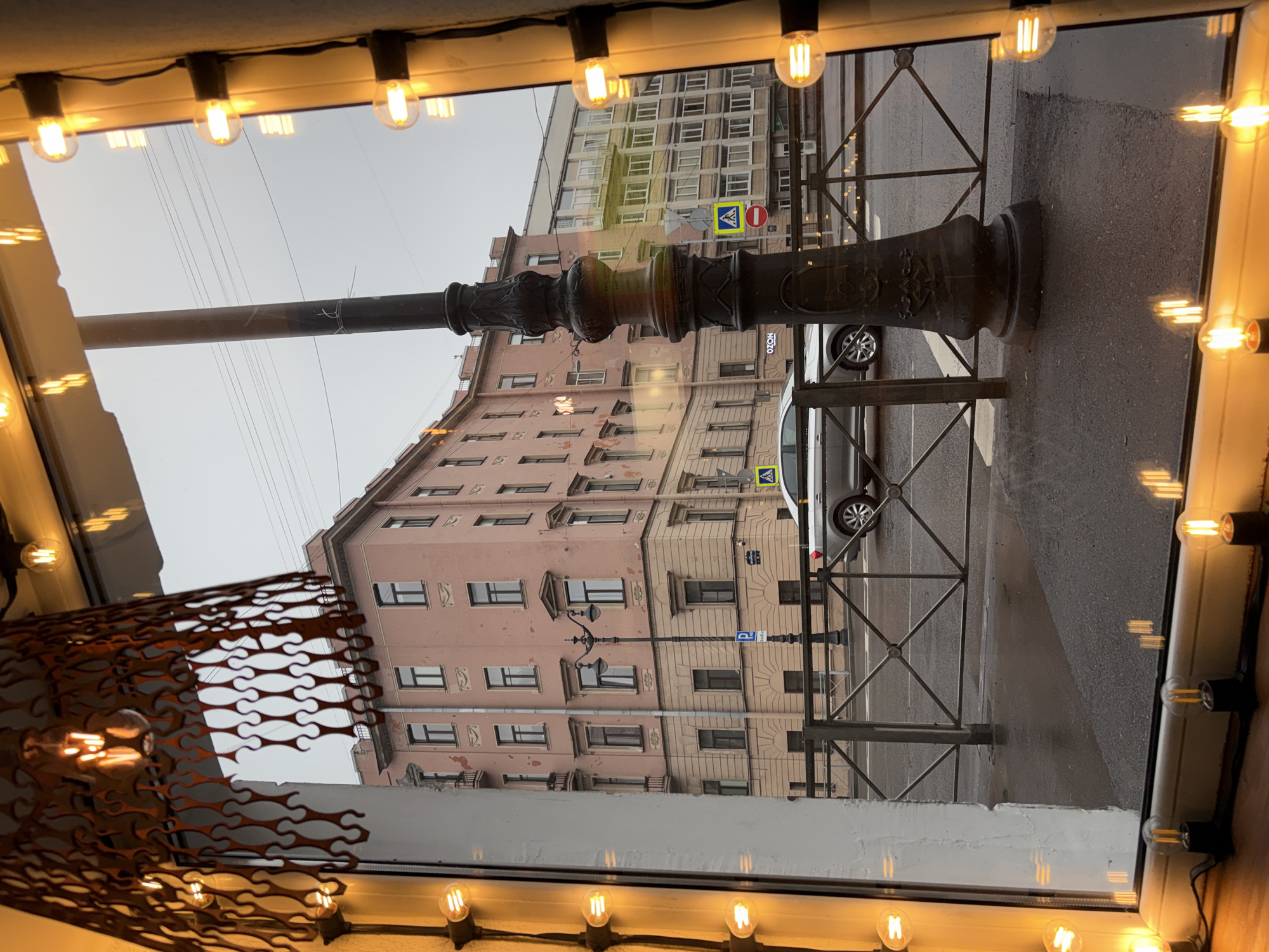 This image depicts a view from indoors, likely through a window, overlooking a street scene in Saint Petersburg, Russia. The window frame is visible, adorned with a string of decorative light bulbs.
The street outside shows buildings with multiple floors and traditional architecture, painted in light pink and beige tones. A street lamp is present, appearing twice in the shot: once partially obscured by the window frame, and another reflected in the glass. Street signs are mounted to the buildings.
A vehicle, possibly a van, is parked on the street. Metal fencing separates the sidewalk from the road. The sky is overcast. The overall scene suggests an urban setting. A decorative piece hangs on the left, partially obscuring the view.