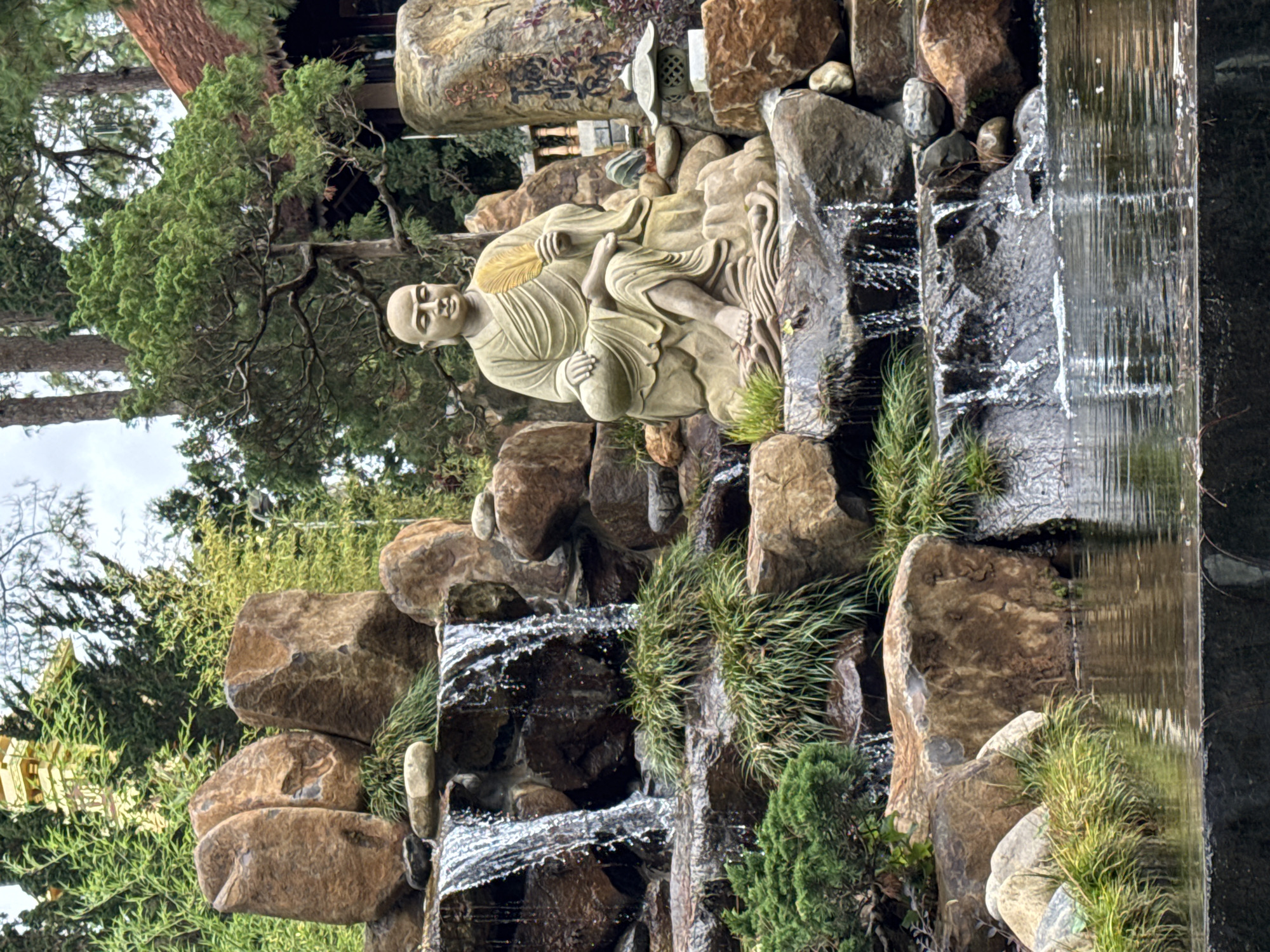 The image displays a serene outdoor scene, likely a landscaped garden or part of a temple complex in Da Lat, Vietnam, featuring a multi-tiered waterfall and a stone statue.
The central subject is a stone statue of a bald, robed figure, resembling a monk or Arhat, seated in a relaxed posture on a rock formation. The figure holds a golden, leaf-shaped fan in its left hand and has a calm, introspective expression. The drapery of its robes is intricately carved, suggesting a sense of movement.
Behind and above the statue, a small, intricate stone lantern is partially visible, set among larger rocks. Further back, verdant green trees with fine needles, possibly pines or junipers, rise against a light sky, contributing to a tranquil backdrop.
To the right of the statue and extending downwards, a dynamic waterfall cascades over multiple levels of dark and light-brown rocks. The water flows smoothly in some sections and creates white, frothy splashes as it tumbles over the rock edges. Various green grasses and small bushy plants grow abundantly around the rocks and along the edges of the water features, adding to the naturalistic feel.
The scene is illuminated by bright, possibly overcast daylight, creating soft shadows and highlighting the textures of the stone, water, and foliage. Some faded or carved characters, possibly Asian script, are faintly visible on a large rock behind the statue, but are not legible. The overall impression is one of peace and contemplation within a carefully designed natural environment.
