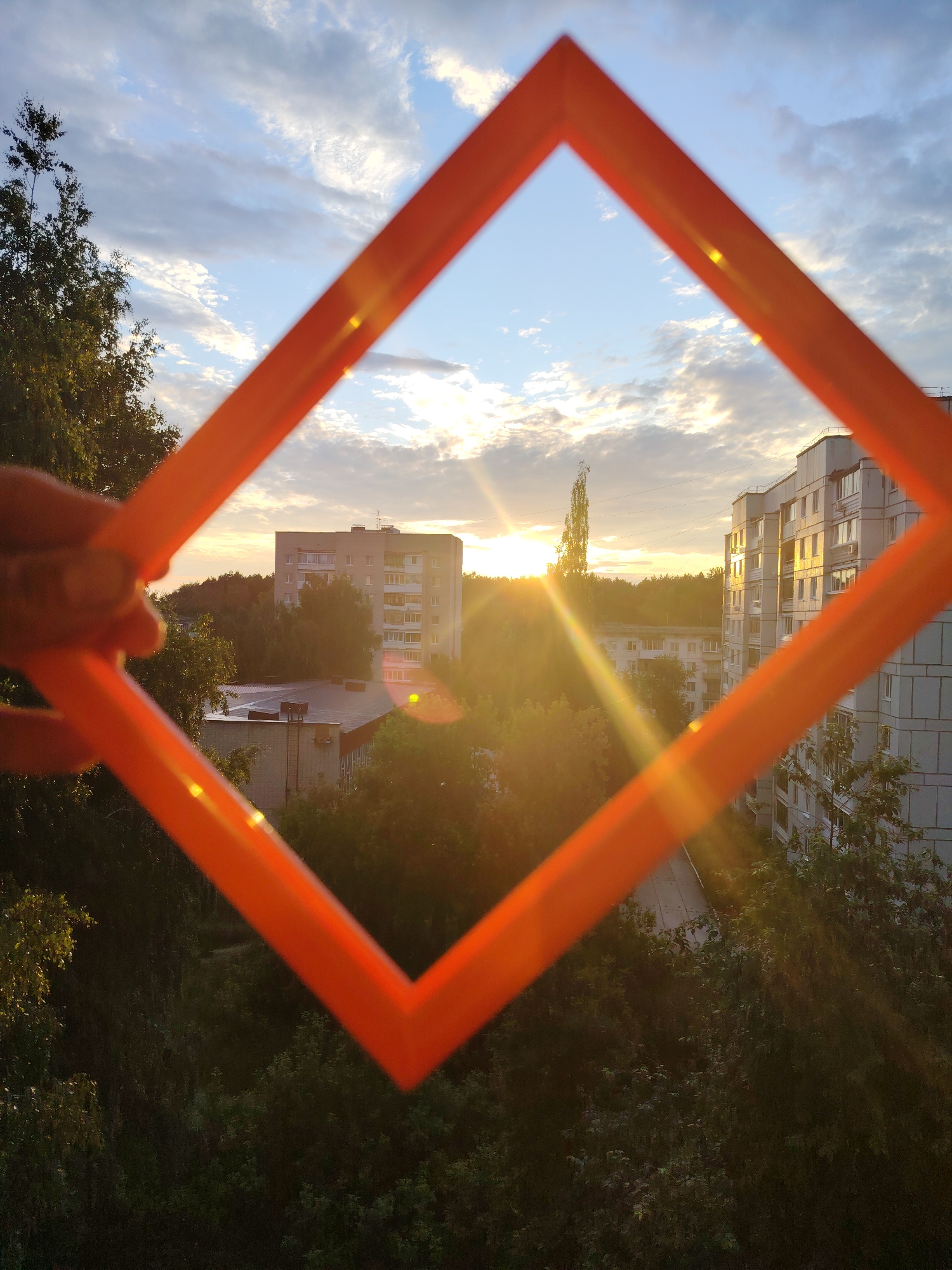 An orange rhombus-shaped frame, held by a person's hand, is positioned in the foreground of a cityscape at sunset. The frame partially obstructs the view of apartment buildings and trees. The sun is visible in the background, casting a warm glow over the scene. The sky is partly cloudy. The location is identified as Chaikovsky, Russia.