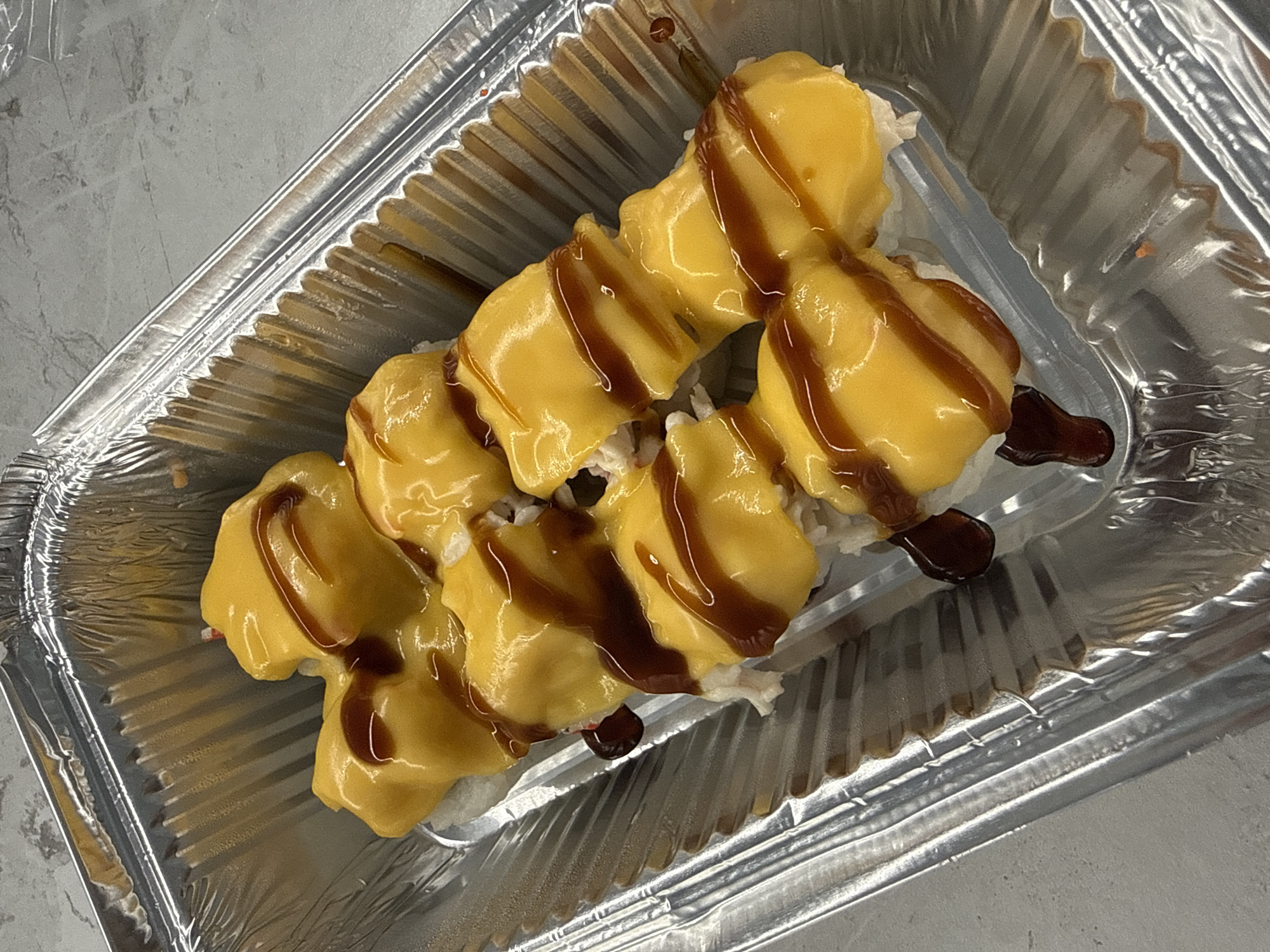 The image shows a take-out container of sushi rolls with a yellow sauce and a brown drizzle on top. The container is made of corrugated aluminum. The background is a light grey surface. This image was likely taken in the city of Lyubertsy, Russia. No people are visible.
