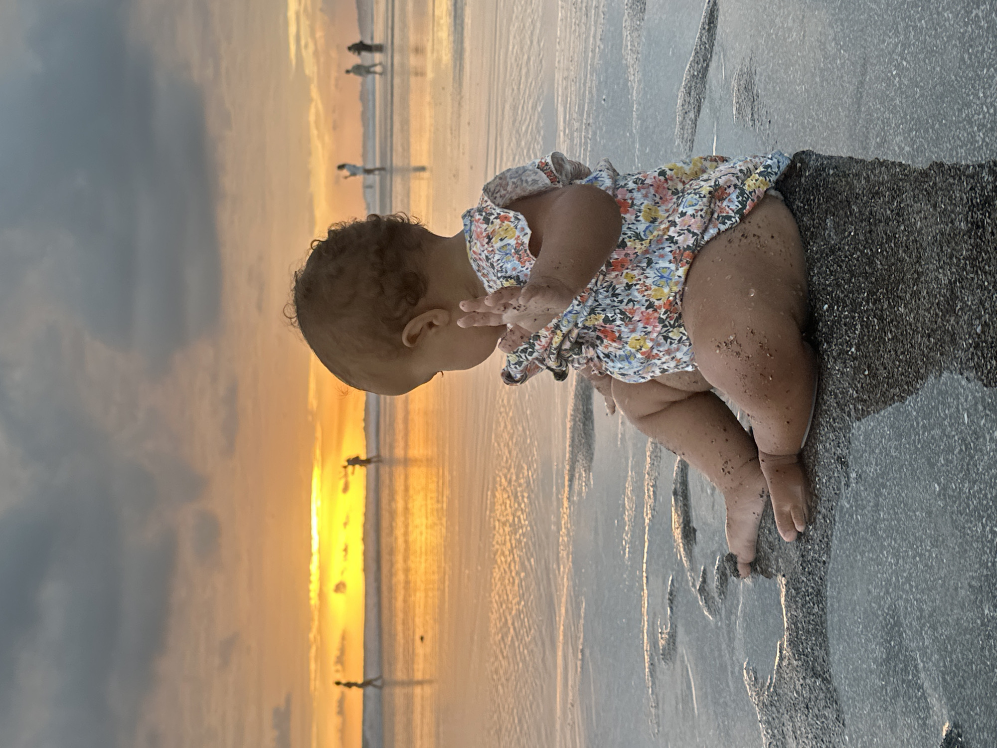 A young child wearing a floral-print romper is sitting on a wet, sandy beach at sunset. The child is facing away from the camera, looking towards the ocean. The sky is filled with dramatic clouds and a bright orange sun is setting on the horizon, casting a reflection on the water. Several silhouettes of people can be seen walking along the shoreline in the distance. The overall scene evokes a serene and peaceful atmosphere. The location is specified as North Kuta, Indonesia.