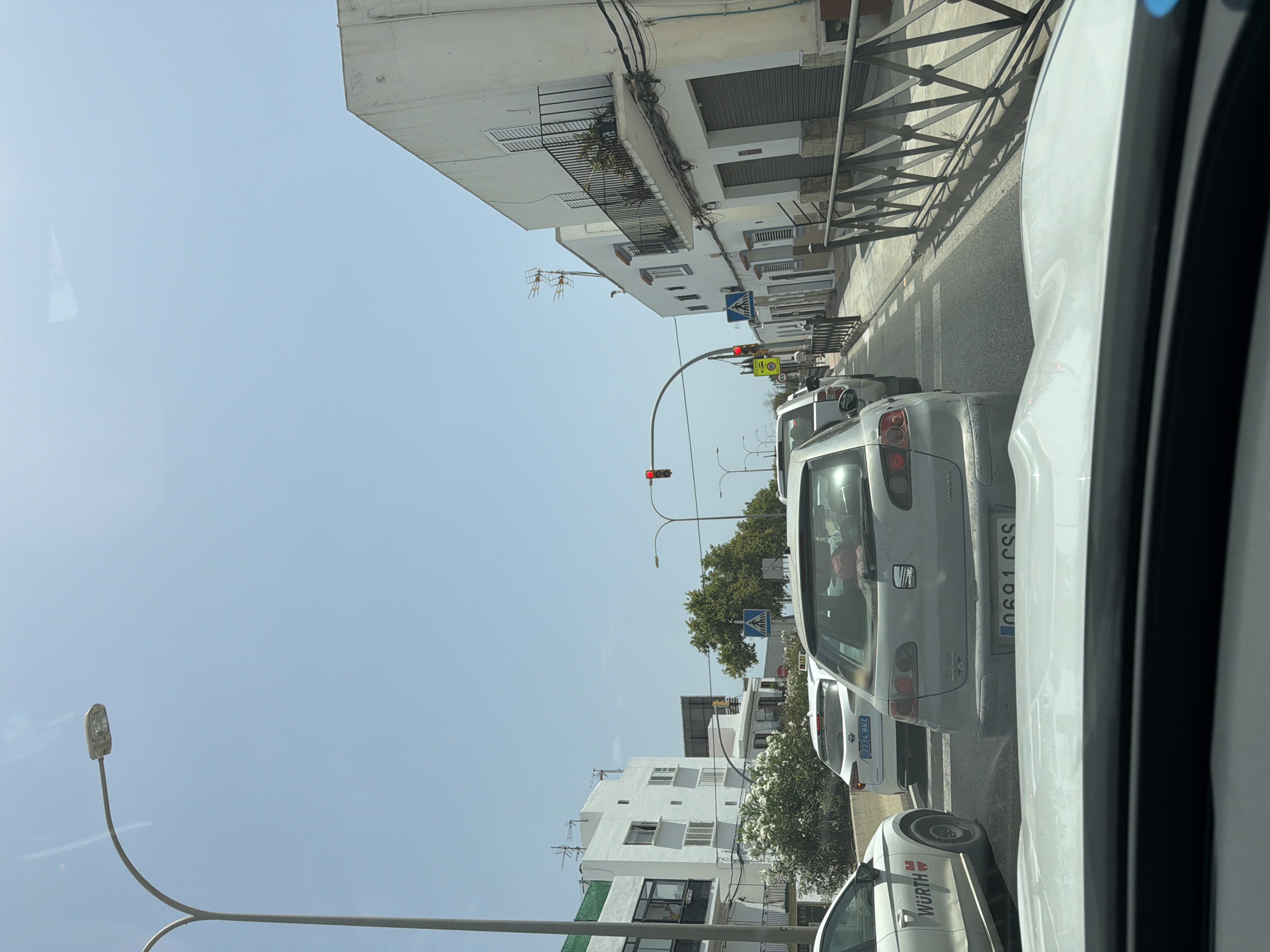 The image depicts a street scene in Can Negre, Spain, on a bright, sunny day. The perspective is from inside a vehicle, looking forward through its windshield, which shows reflections. The street is lined with white buildings, some with balconies and potted plants. There is traffic congestion, with several cars stopped in a lane. A white car with the "WURTH" logo is visible on the left. A light gray hatchback with the license plate "0691 CSS" is directly in front, and a white car with license plate "2334 MMX" is ahead of that. Traffic signals, including a red light, are visible. Pedestrian crossing signs are also present. Trees and vegetation are present along the street.