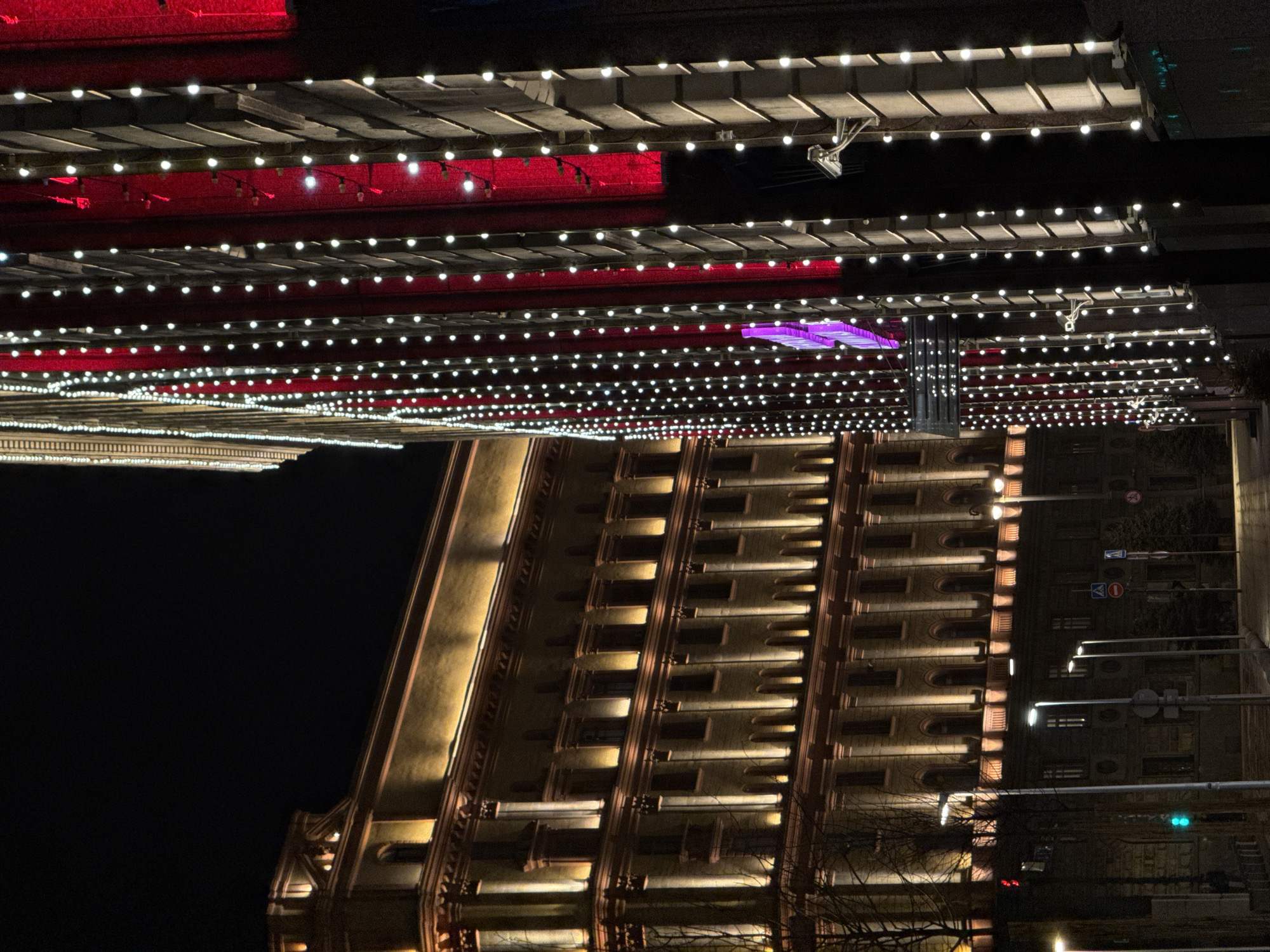 Here is a description of the image:
The image depicts an upside-down view of a building illuminated at night in Moscow, Russia. String lights adorn structures above the building. The lights are strung on several horizontal planes, creating a visually layered effect. The building itself is multiple stories tall with evenly spaced window features and architectural details highlighted by vertical lighting. The environment is dark, with the building and decorative lights providing the main sources of illumination. Street signs and trees are at the edge of the building.