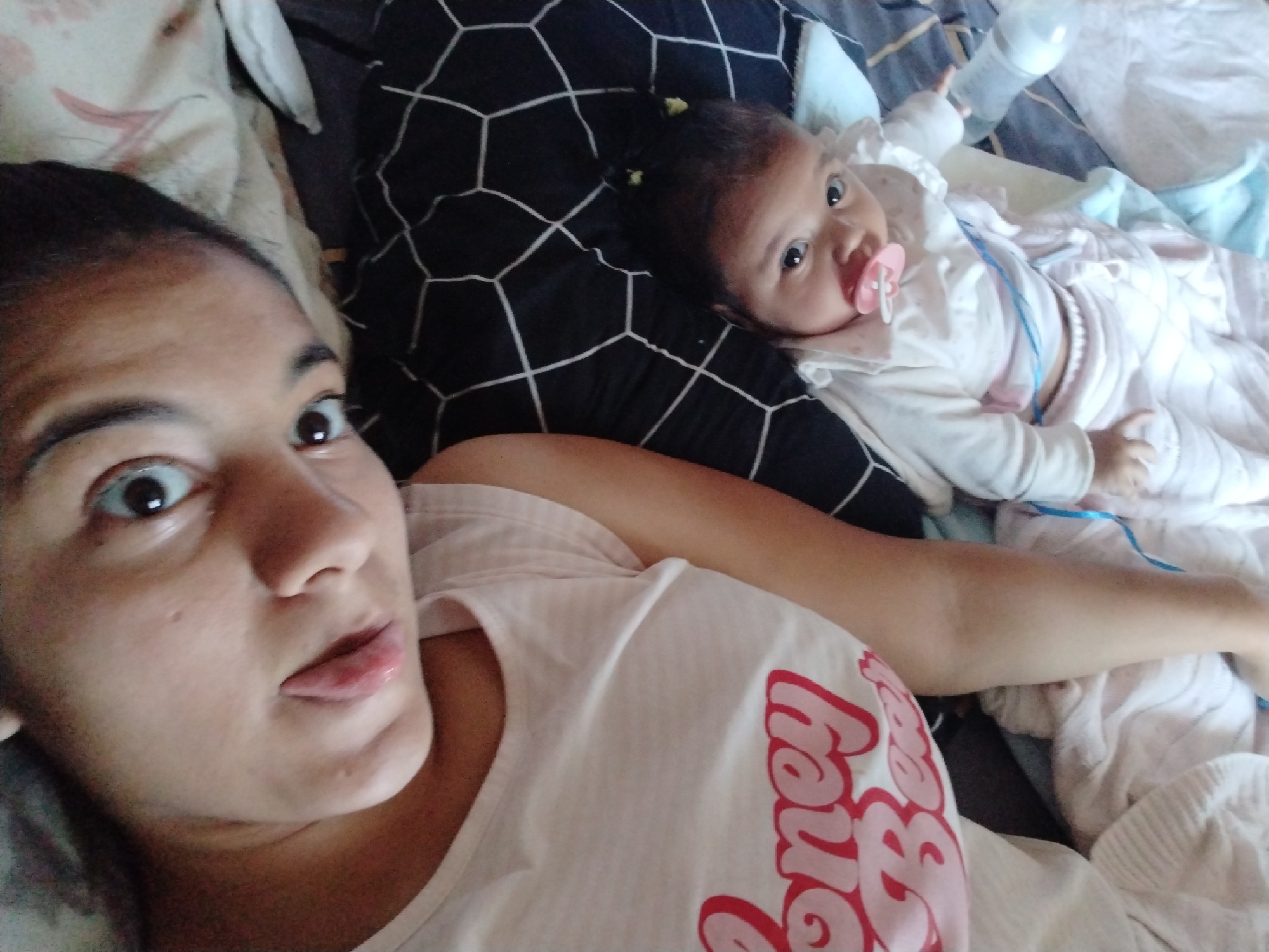 A woman and an infant are lying on a bed in an indoor setting. The woman is in the foreground, facing the camera with a slight sideways glance. She has dark hair pulled back from her face and is wearing a white t-shirt with pink lettering. The infant lies behind her, on a pillow with a black and white geometric pattern. The infant is also looking at the camera, with a pacifier in its mouth. The infant is dressed in white clothing with a blue string visible. The background includes bedding in shades of blue and white, and a baby bottle. The location is Unknown, Unknown.
