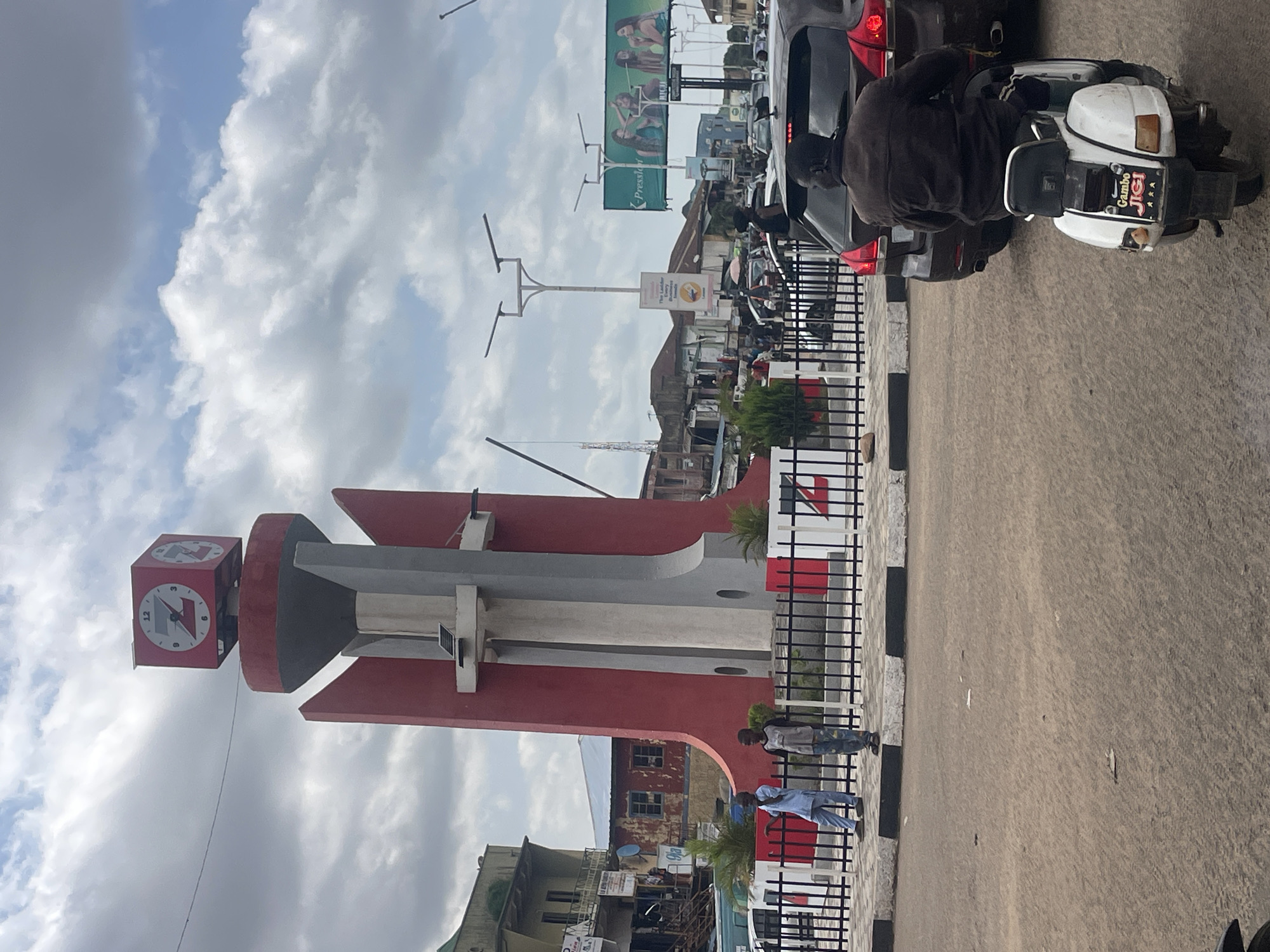 The image depicts the Jos Main Market Clock Tower, a prominent landmark in Jos, Nigeria. In the foreground, a person is riding a white scooter with the inscription "Gambo Jigi" on the back. The scooter is in traffic, with several cars behind it. The clock tower, painted red and gray, is visible in the mid-ground, topped with a red clock. Two young boys stand near a black fence in front of the tower. In the background, a billboard advertising "K-Presslor" and "RICHI" is visible, along with buildings and streetlights, suggesting a busy urban environment. The sky is partly cloudy.