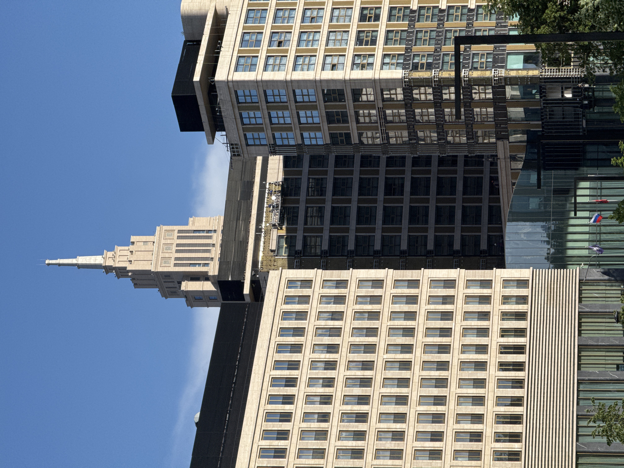The image showcases several buildings in Moscow, Russia, under a clear blue sky. The architectural styles vary, with one building featuring a slender, stepped tower, possibly reminiscent of Stalinist architecture. The other buildings are more modern with grid-like facades of many windows. Some structures have reflective surfaces. Russian flags are visible on one of the buildings. There is greenery, which could include trees, visible on the right side of the image.
