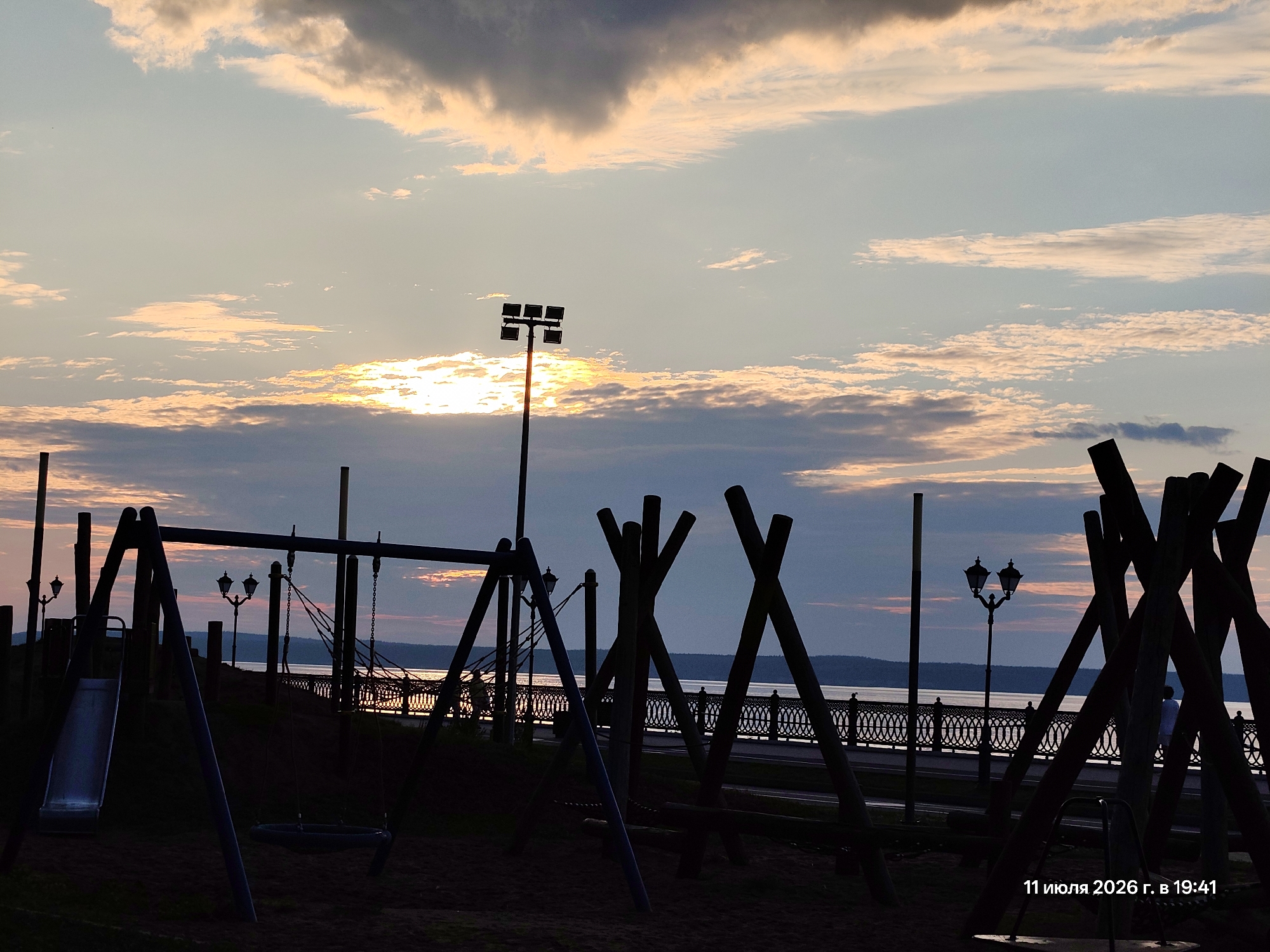 The image shows a playground in silhouette against a sunset sky. The sky is filled with clouds, with the sun peeking through in the upper left portion of the image. In the foreground, there are various playground structures, including swings and a slide. In the background, there is a body of water, a decorative fence along the water, and trees or hills on the opposite shore. Two streetlights are visible, one taller with multiple bulbs and another with two traditional lanterns. A date and time stamp, "11 июля 2026 г. в 19:41", is visible in the bottom right corner. There are a few indistinct figures walking along the waterfront promenade. The overall scene depicts a peaceful evening by the water.