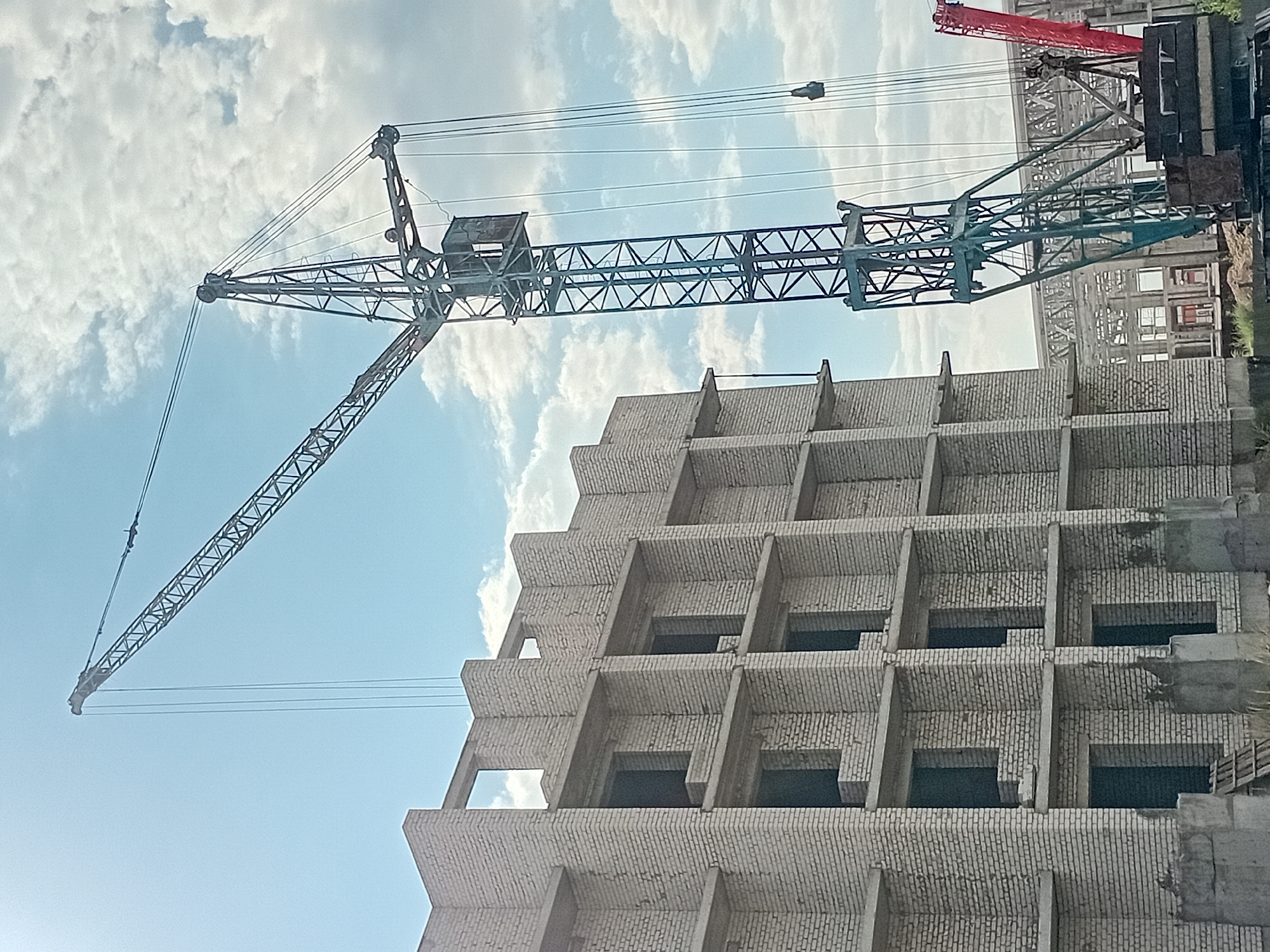 A construction site in Unknown, Unknown shows a partially completed multi-story building made of white brick. Windows and doorways are visible on the unfinished structure. A tall, dark green construction crane with a long boom extends over the building, its cables reaching towards the top. The sky is blue with scattered white clouds. In the background, another red crane is partially visible, and a skeletal metal structure of another building is under construction. The ground appears to be dirt with some debris.