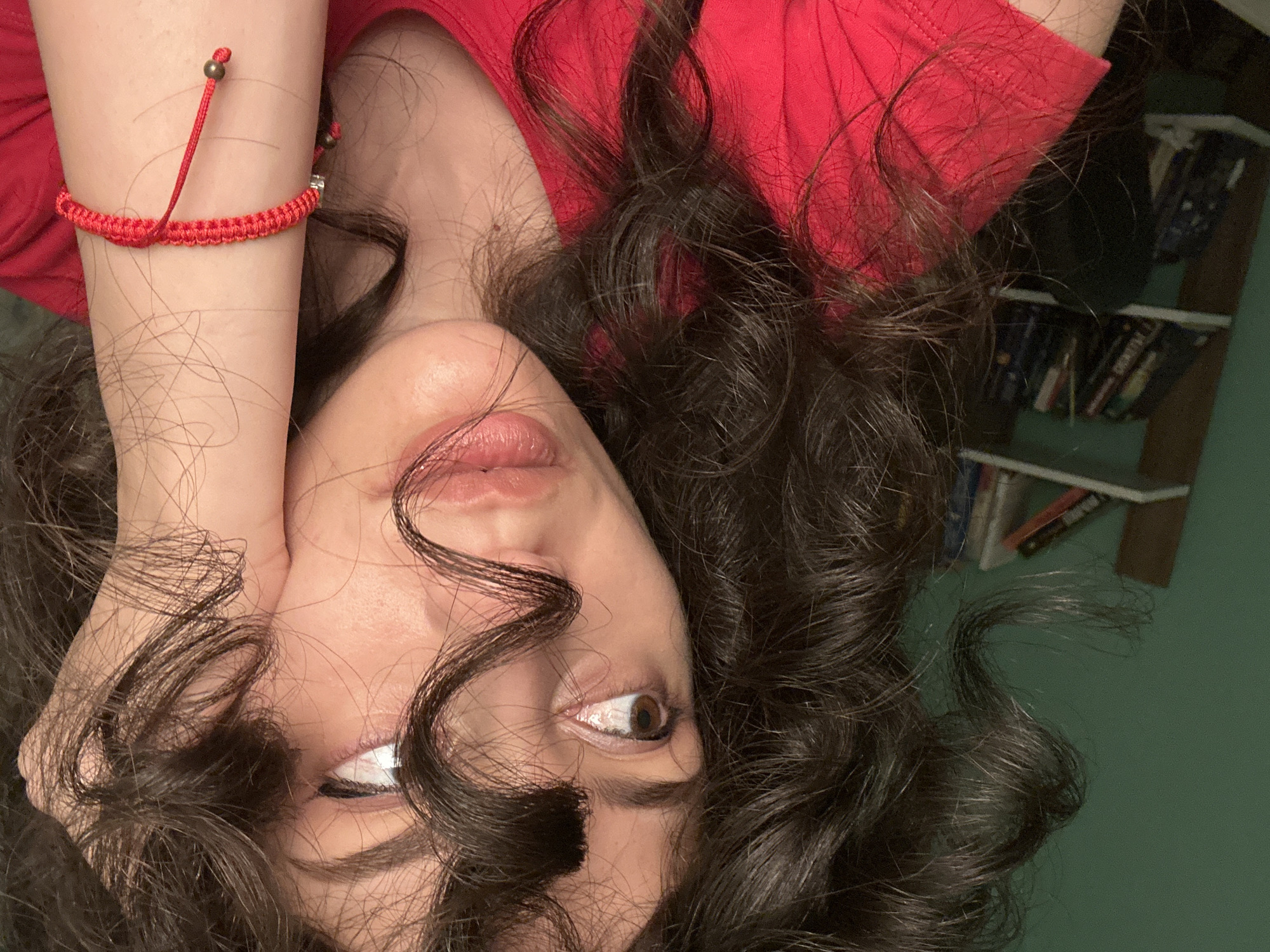 The image shows a close-up of a woman's face and upper body. She has dark, curly hair that frames her face, and her left hand is resting on her cheek. She is wearing a red t-shirt and a red bracelet on her left wrist. In the background, there is a green wall and a bookshelf filled with books. The scene appears to be indoors, possibly in a bedroom or living room. There is no indication of the specific location within the city of Sofia, Bulgaria.