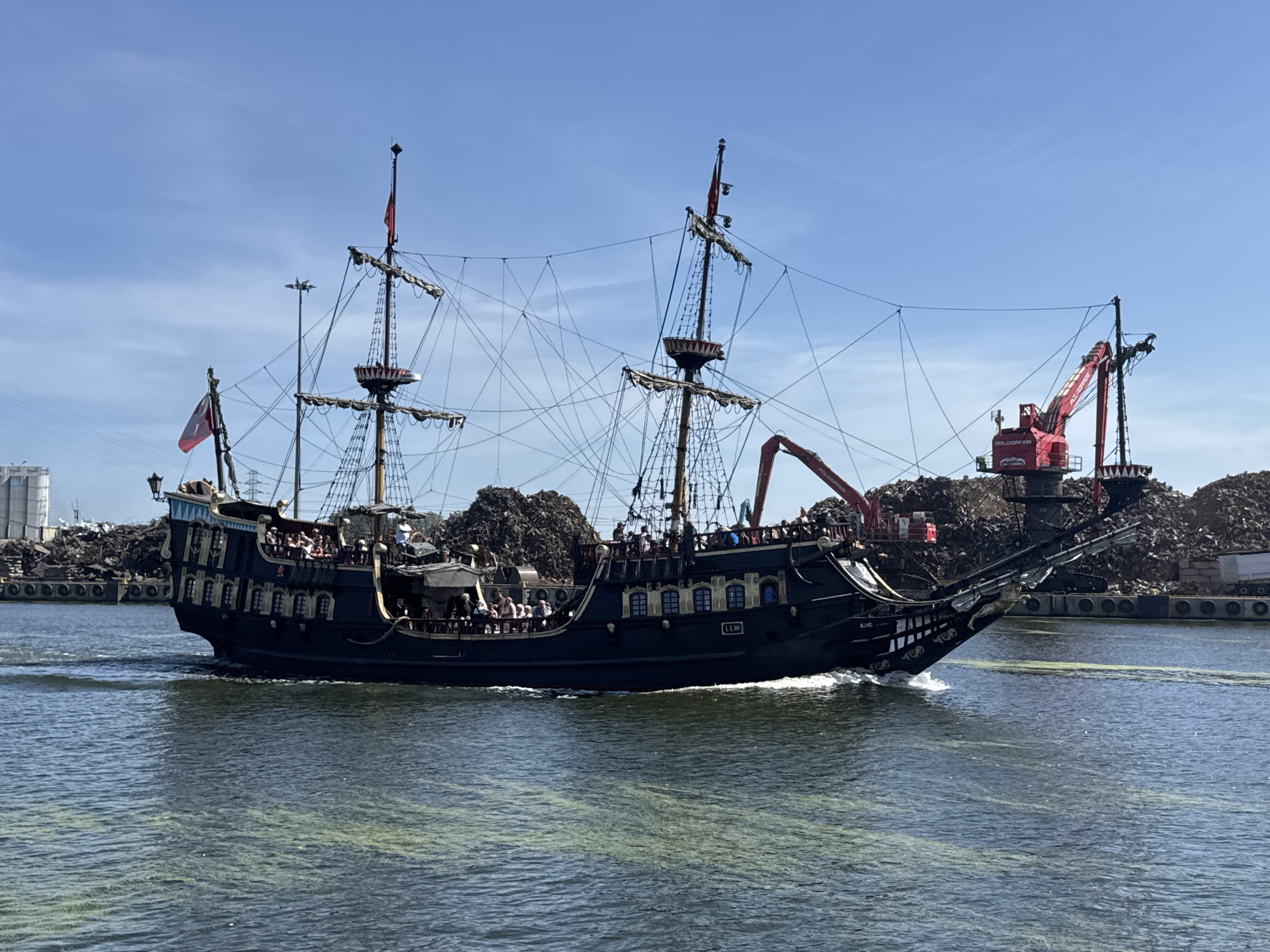 A black, three-masted sailing ship, resembling a pirate ship, is sailing on a body of water. The ship has multiple decks with people visible on them. Red flags are flying from the masts, and intricate rigging is present. The ship's bow features a decorative figurehead and elaborate carvings.
In the background, a large pile of what appears to be scrap metal or debris is visible, along with industrial structures and cranes, one of which is red. The scene suggests an industrial or port area. The sky is clear and blue with some wispy clouds, indicating fair weather. The time of day appears to be daytime, likely sunny. The water has a greenish-yellow hue in patches, possibly due to algae or pollutants.
The ship's name "LEW" is visible on the side of the hull. The overall atmosphere seems calm and perhaps a bit unusual, given the juxtaposition of the historical-style ship with the industrial backdrop. The people on the ship appear to be tourists or passengers enjoying the excursion. The location is identified as Gdańsk, Poland.