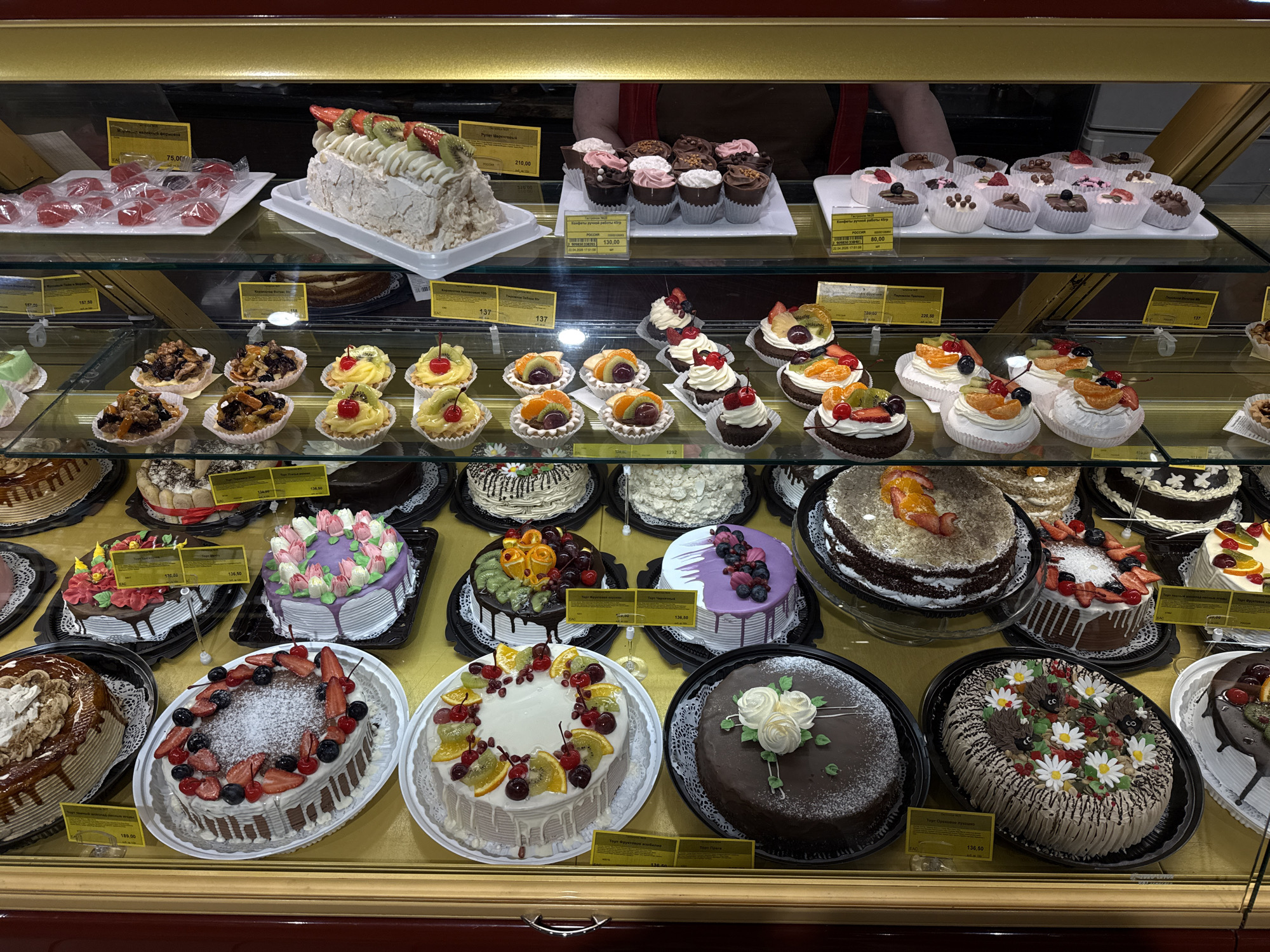 This image captures a vibrant display case filled with a wide variety of desserts and pastries, likely in a bakery or confectionery shop. The scene is brightly lit, highlighting the colorful creations behind clear glass shelves.
On the top shelf to the left, there's a tray of individually wrapped red jelly candies, and next to it, a large meringue cake adorned with sliced strawberries and kiwi. On the right side of the top shelf, a white tray holds numerous small cupcakes or chocolate cups with pink or white frosting and sprinkles. A person's arm and torso are partially visible in the background, suggesting staff or a customer.
The middle shelf presents an array of smaller individual desserts. To the left, there are tarts filled with nuts and dried fruits. To the right, small individual meringue or cream desserts are topped with fresh fruits like kiwi, tangerine segments, and cherries.
The bottom shelf showcases an assortment of larger cakes. Notable examples include a round cake with white chocolate drizzle, powdered sugar, and a generous topping of strawberries, blueberries, and cherries. Another round white cake features a colorful garland of kiwi, tangerine, grapes, and cherries. A dark chocolate cake is elegantly decorated with white frosting roses. Further to the right, an oval cake depicts a whimsical scene with hedgehog figures, white daisies, and berries. Other cakes with various frostings and decorations, including floral patterns and fruit toppings, are also visible.
Numerous yellow price tags written in Cyrillic script are scattered throughout the display. Visible text includes prices such as "157,50", "189,00", "75,00", "136,50", "210,00", "137", "130,00", "80,00", and "220,50". Some labels also read "Конфеты ручной работы 45гр" (Handmade candies 45gr), "РОССИЯ" (RUSSIA), and a date/timestamp "22.04.2006 17:51:00". The overall impression is one of abundance and careful presentation, inviting customers to purchase these sweet treats.