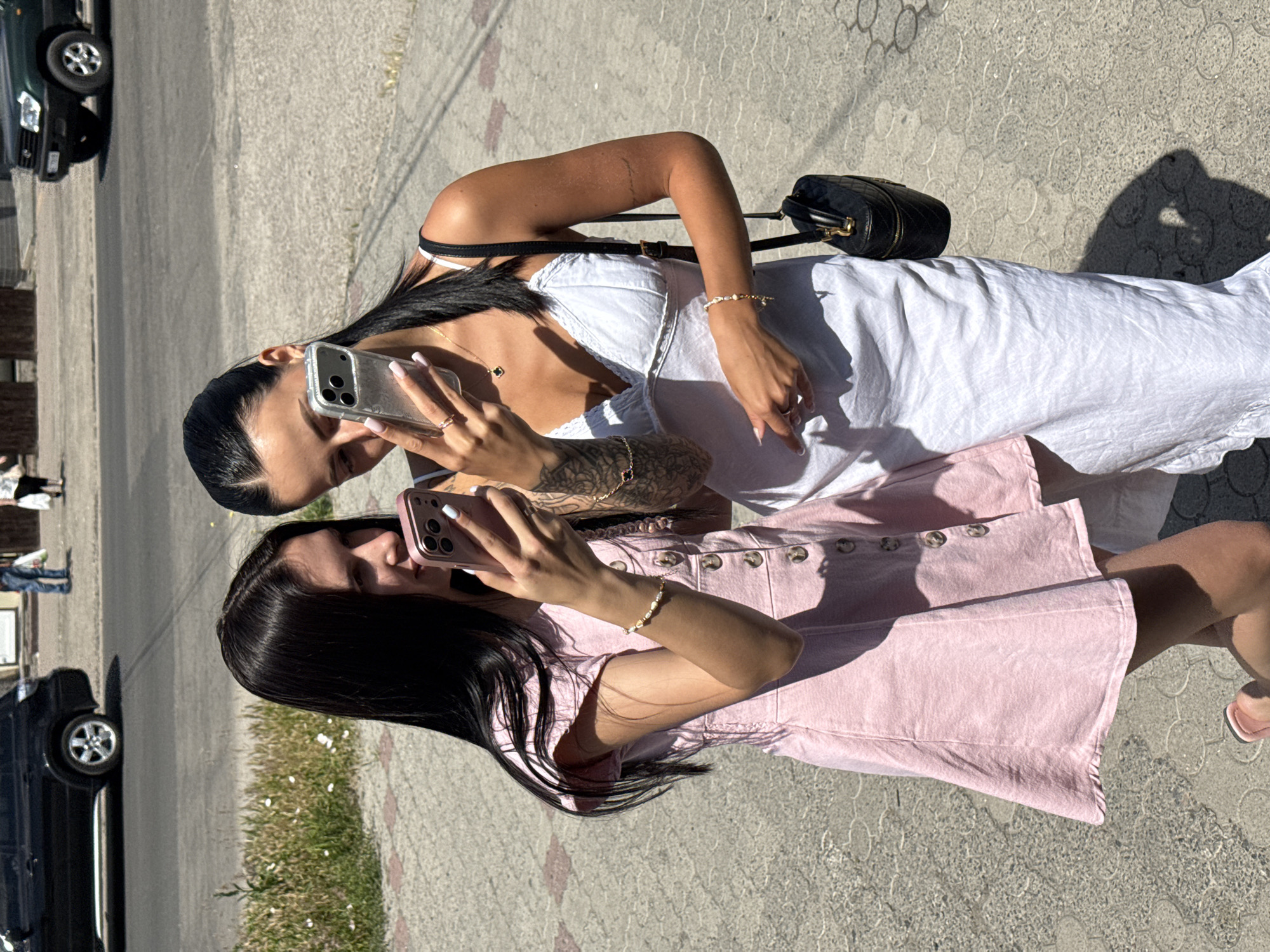 Two young women are standing on a sidewalk, holding smartphones and looking at them. The woman on the left is wearing a pink dress and the woman on the right is wearing a white dress. They are both facing the camera. The background shows a street with cars and buildings. The location is identified as Unknown, Unknown.
